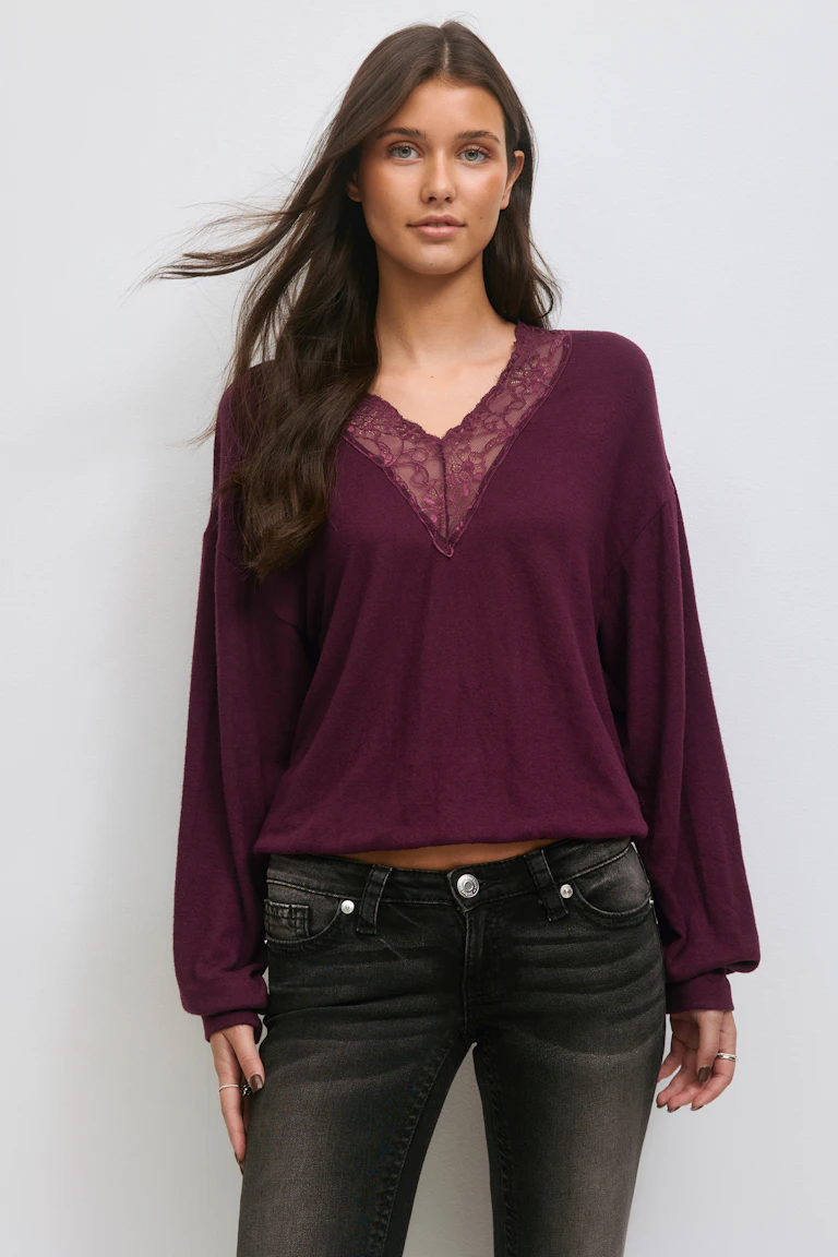 Luumuinen pitkähihainen V-pääntiepusero, jossa on pitsipanos ja rennot pallohihat, yhdistettynä haalistuneisiin mustiin farkkuihin.