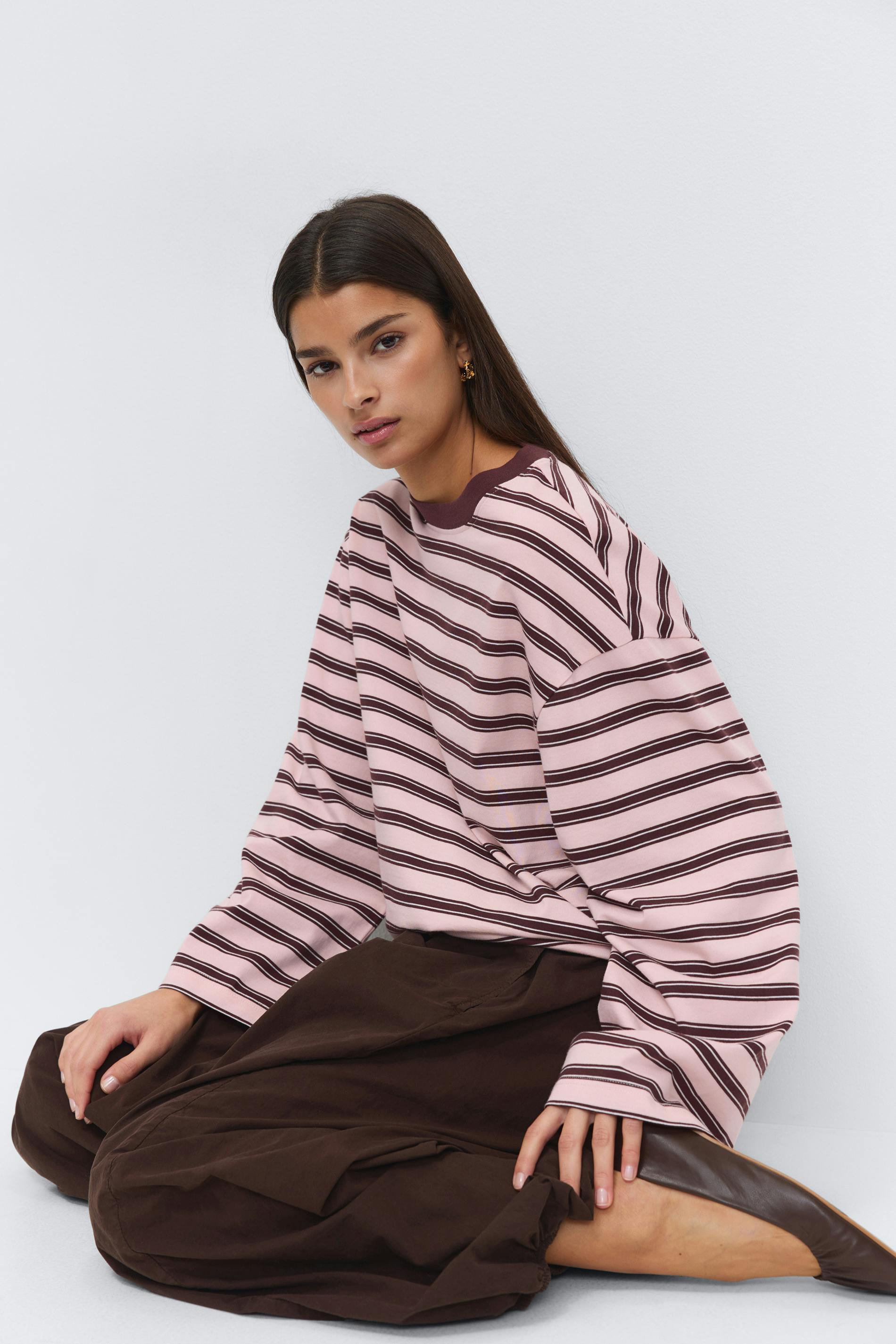 Roze en bruine horizontaal gestreepte oversized top met lange mouwen en een donkerbruine ronde hals, losse donkerbruine broek met wijde pijpen, bruine leren flats en kleine gouden oorbellen.