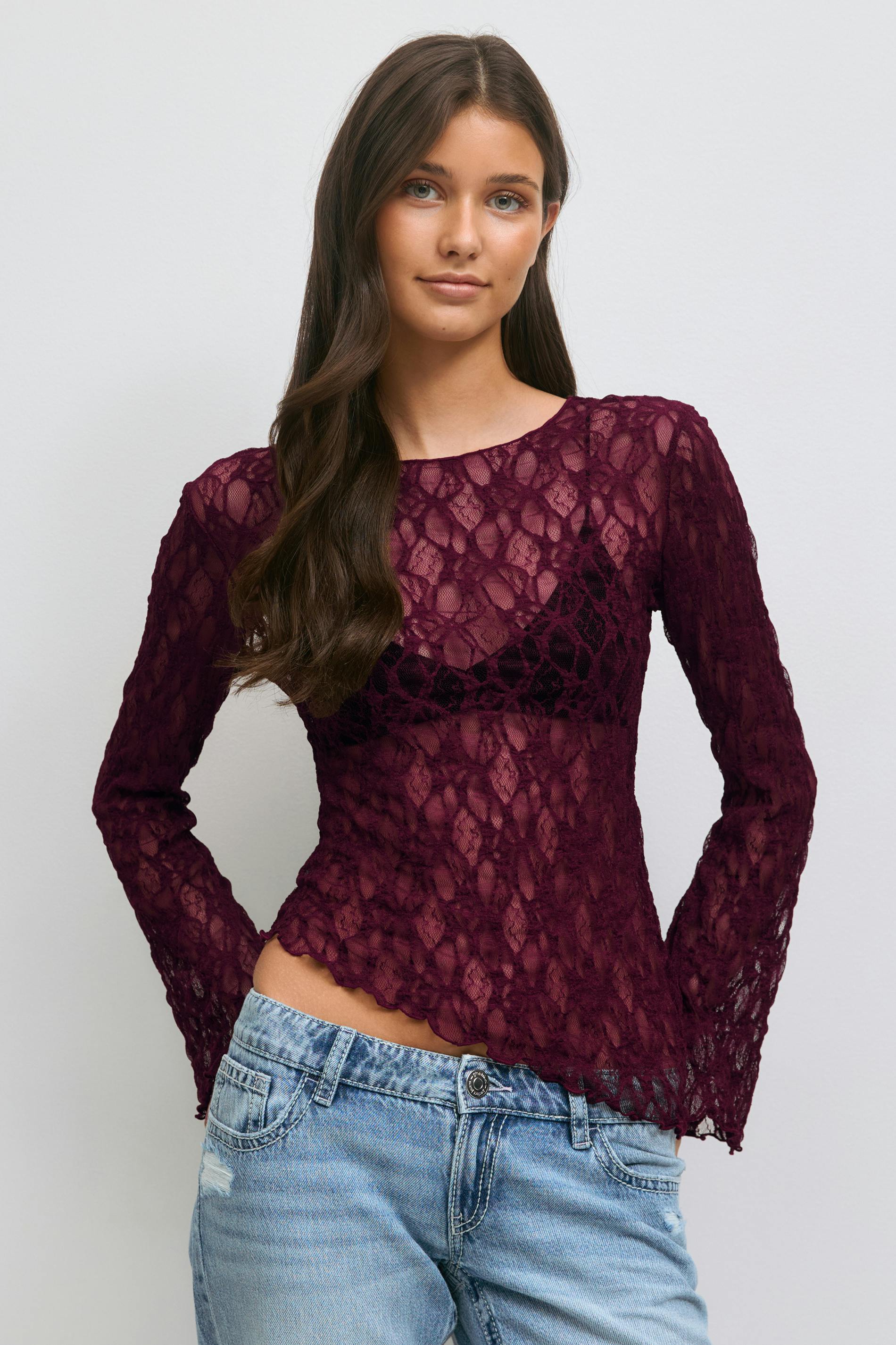 Sheer bordeauxrode kanten top met lange mouwen en geschulpte zoom, gedragen over een zwarte bralette, gecombineerd met een laag uitgesneden denimjeans.