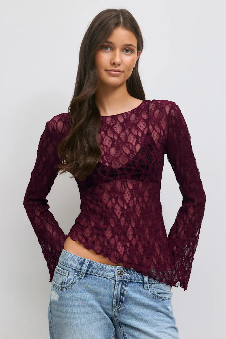 Sheer bordeauxrode kanten top met lange mouwen en geschulpte zoom, gedragen over een zwarte bralette, gecombineerd met een laag uitgesneden denimjeans.