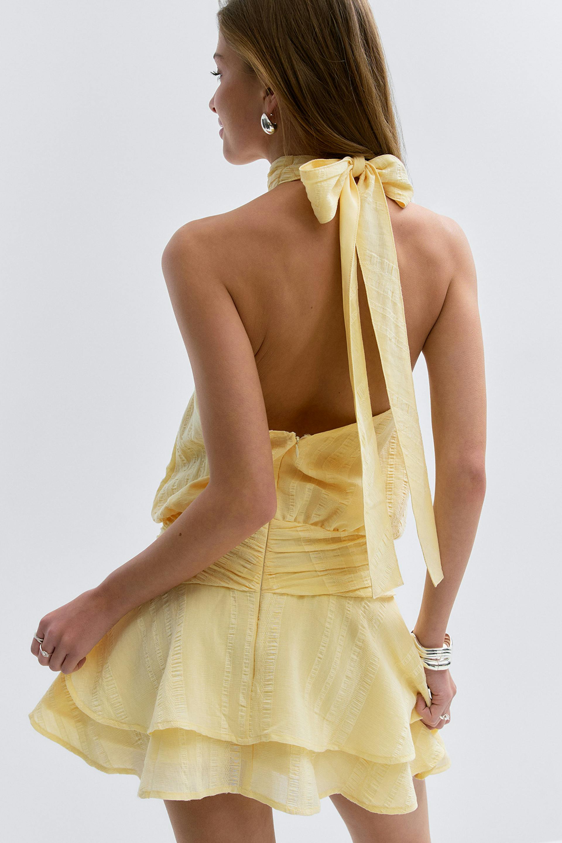 Mini robe dos nu texturée jaune pâle avec un nœud noué sur la nuque, dos ouvert, taille froncée et jupe volantée à étages ; associée à des boucles d'oreilles argentées, des bracelets superposés et des bagues.