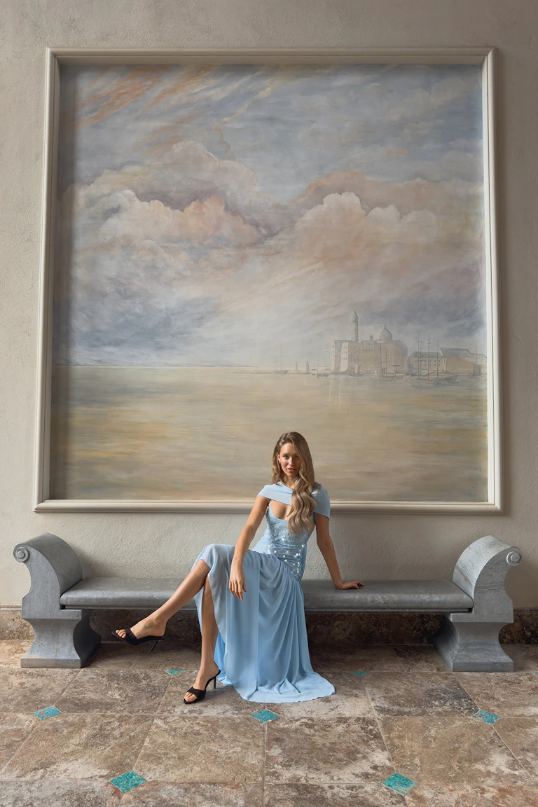 Lyseblå, gulvlang aftenkjole med asymmetrisk off-shoulder-utringning, paljettbesatt, figursydd overdel, lårhøy glidelås og bølgende skjørt i chiffon, kombinert med svarte, høyhælte muldyr med åpen tå.