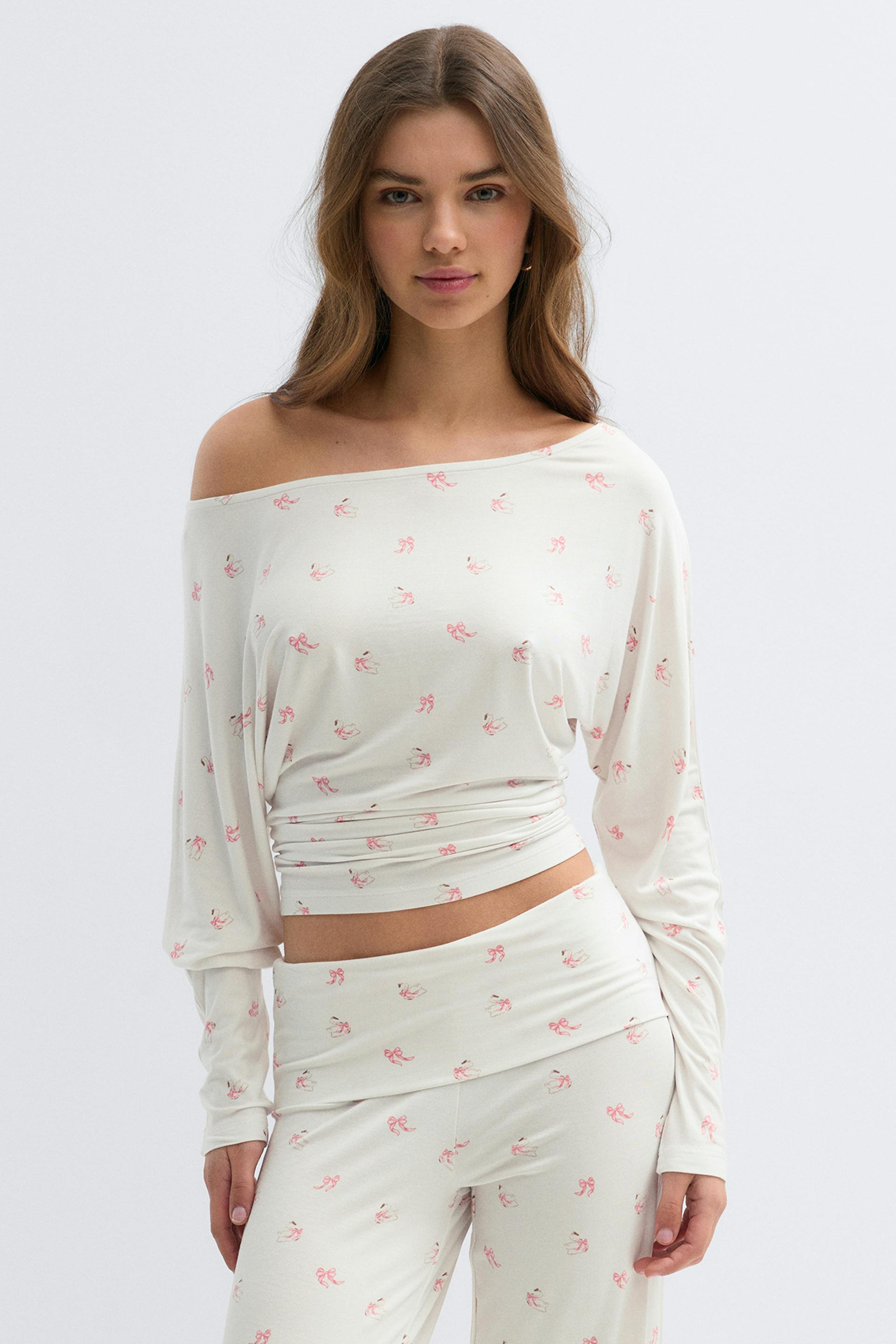 Valkoinen, olkapäätön, pitkähihainen, leikattu toppi, jossa on röyhelöinen vyötärönauha ja kauttaaltaan pieni vaaleanpunainen rusettipainatus, yhdistettynä yhteensopiviin korkeavyötäröisiin, rentoihin housuihin.