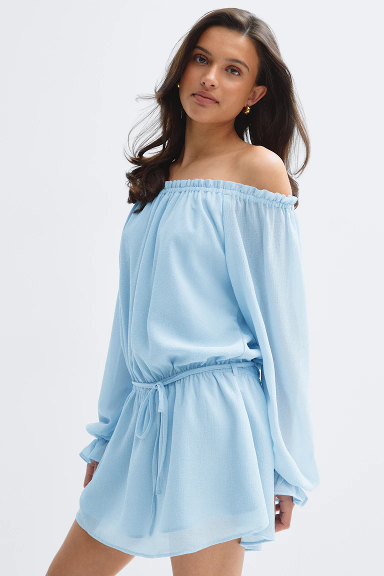 Off-the-shoulder light-blue long-sleeve mini dress with an elasticized ruffled neckline, cinched drawstring waist and flowing skirt, paired with small gold hoop earrings.