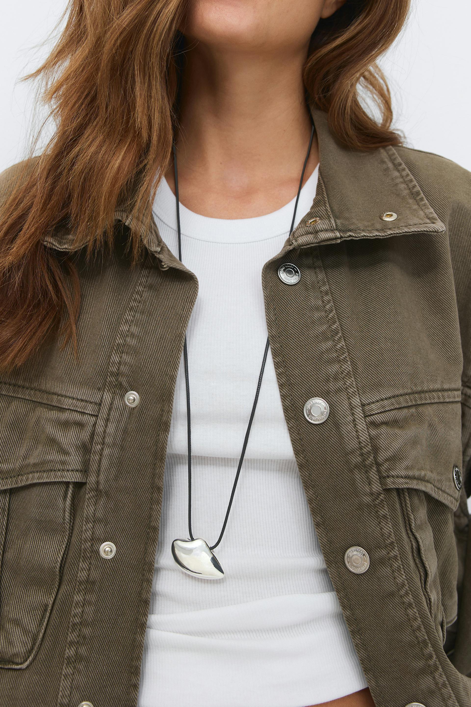 Olive-green utility jacket with metal snap buttons worn over a white ribbed crew-neck crop top, styled with a long black cord necklace bearing a silver abstract pendant.