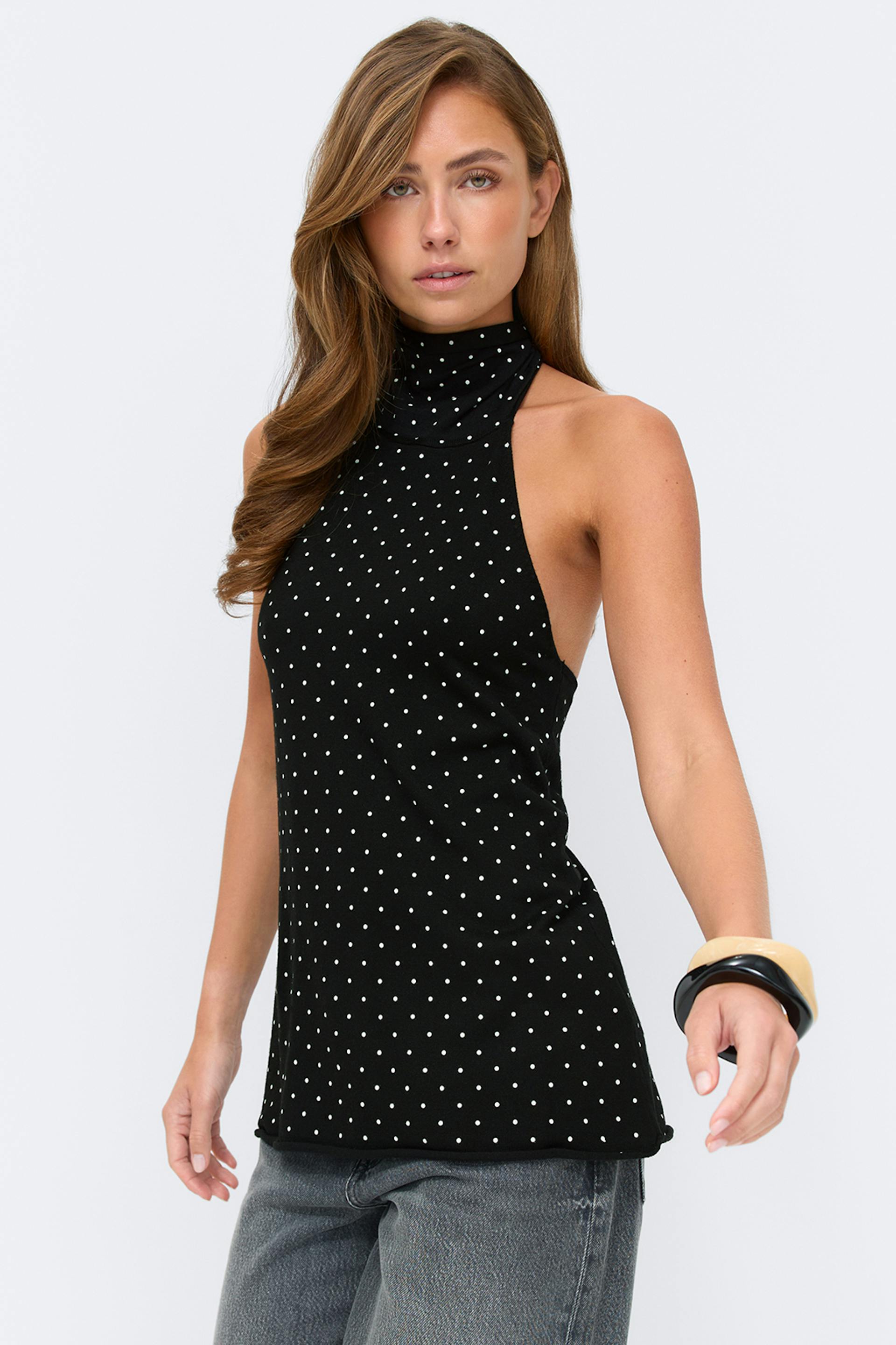 Musta hihaton, korkeakaulainen halter-toppi, jossa on pieniä valkoisia pilkkuja, yhdistettynä haalistuneisiin harmaisiin farkkuihin ja pinottuihin mustiin ja beigeihin rannerenkaisiin.