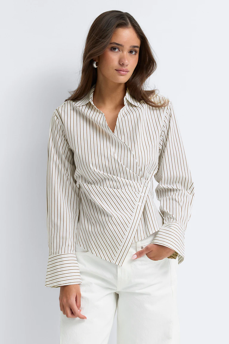 Creme-beigefarbene, vertikal gestreifte Wickelbluse mit asymmetrischer Knopfleiste und breiten Manschetten, kombiniert mit weißen High-Waist-Jeans und kleinen silbernen Ohrringen.