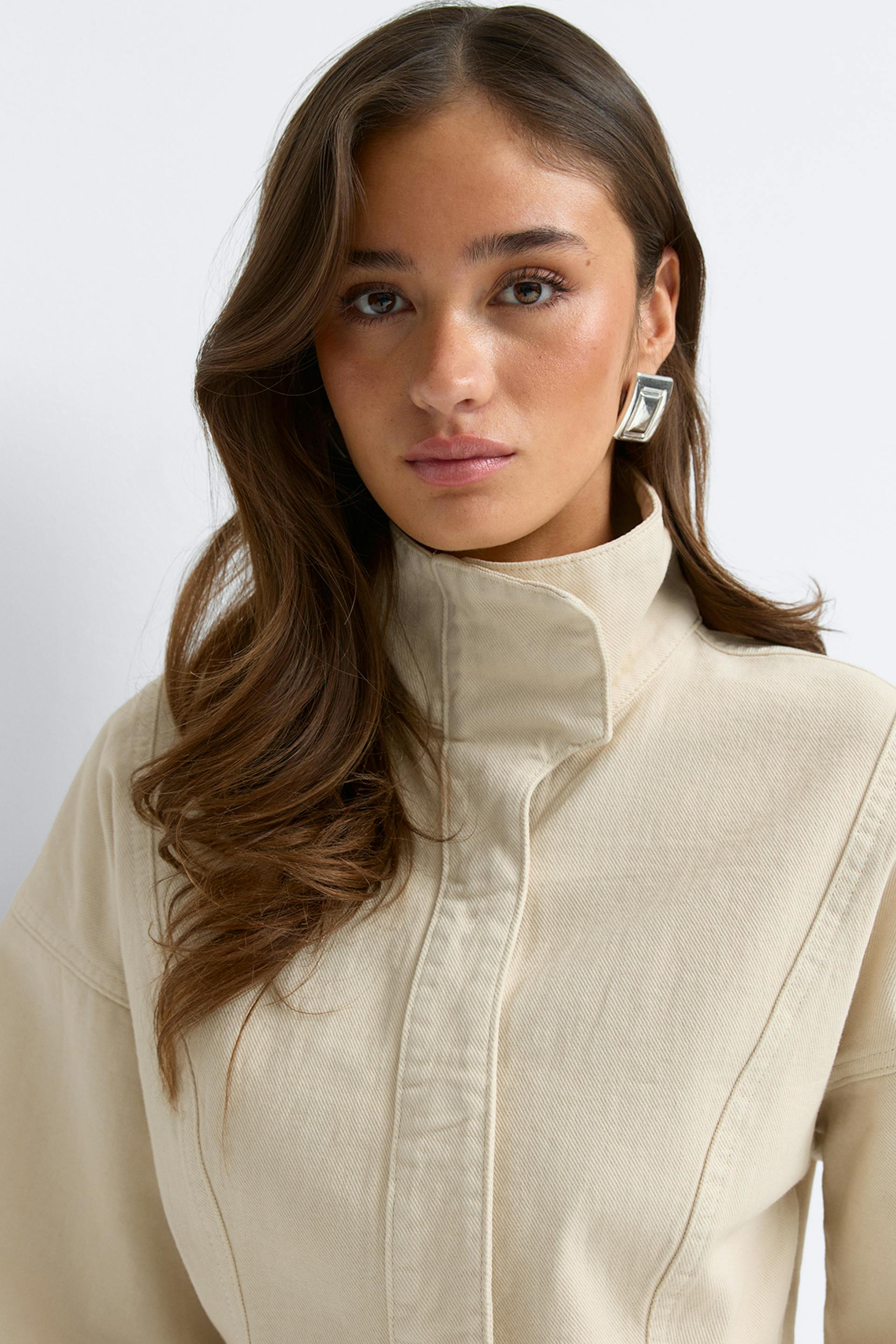 Beige korkeakauluksinen strukturoitu takki, jossa on piilotettu etukansi ja pitkät hihat, yhdistettynä kulmikkaaseen hopeiseen statement-korvakoruun.
