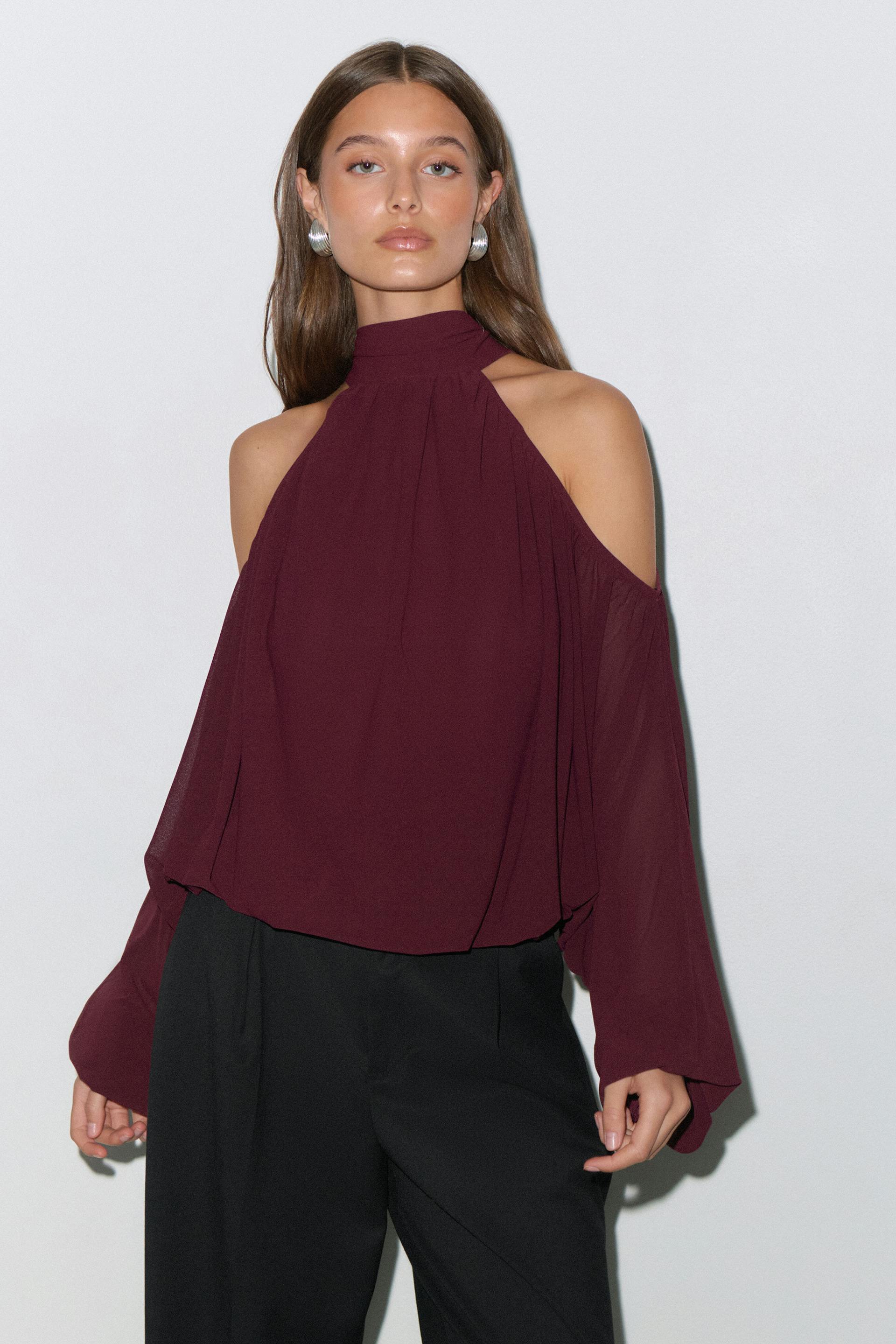 Model wearing a deep burgundy high-neck cold-shoulder blouse with long billowy sleeves, paired with black tailored trousers and silver hoop earrings.