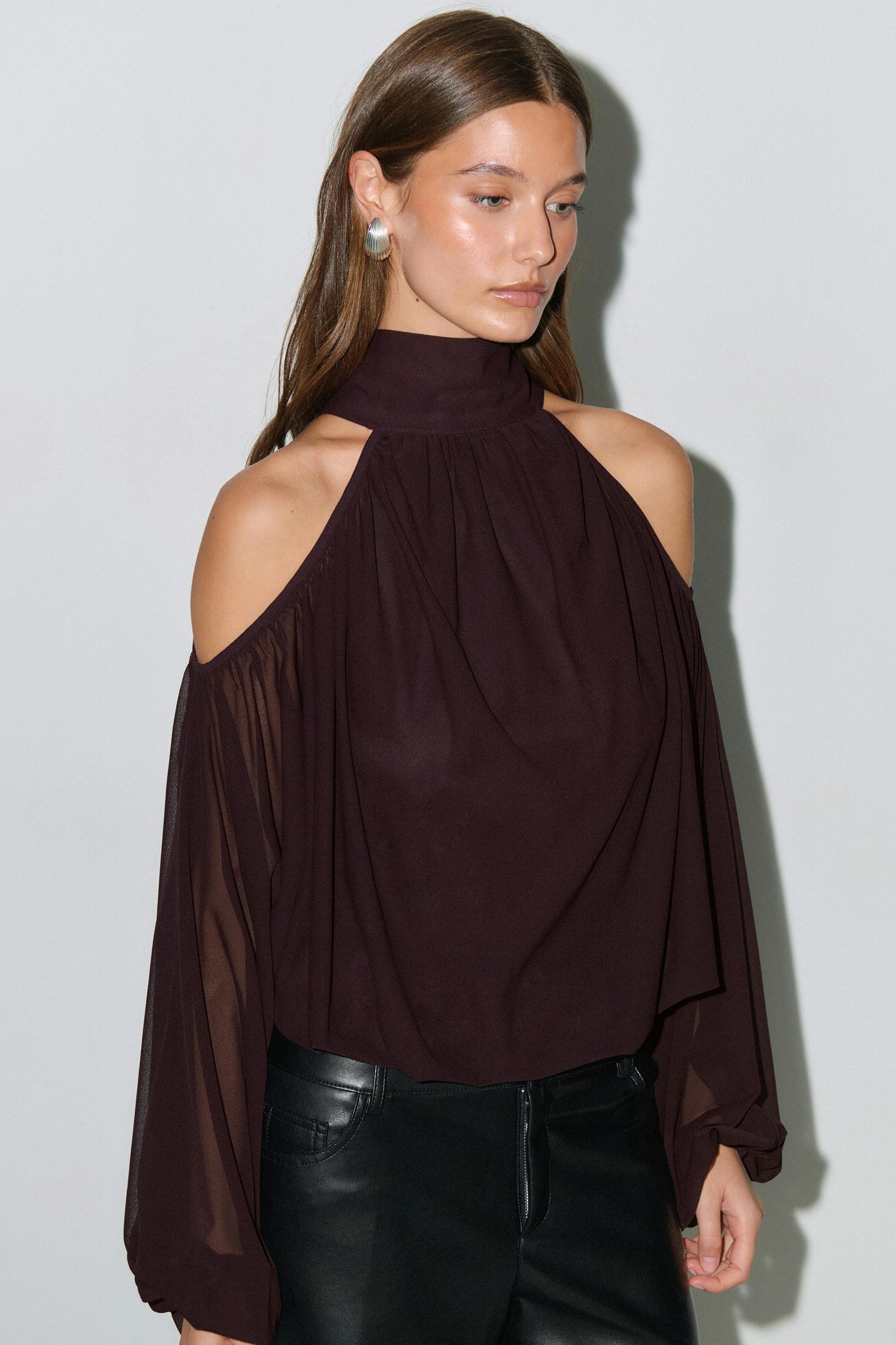 Model wearing a high-neck, cold-shoulder deep plum blouse with gathered yoke and sheer long bishop sleeves, paired with black leather trousers.