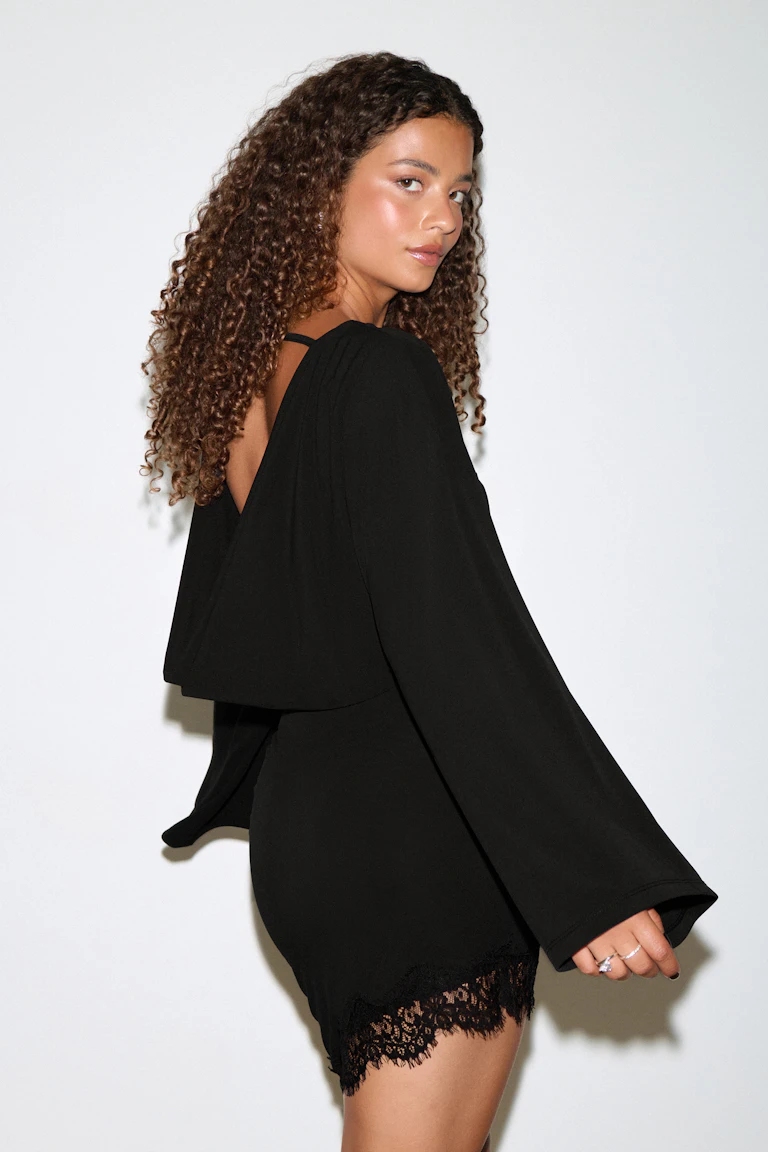 Schwarzes Minikleid mit tailliertem Rock, langen, ausgestellten Ärmeln, drapiertem, offenem Rücken mit dünnem Schulterriemen und Spitzenbordüre am Saum.