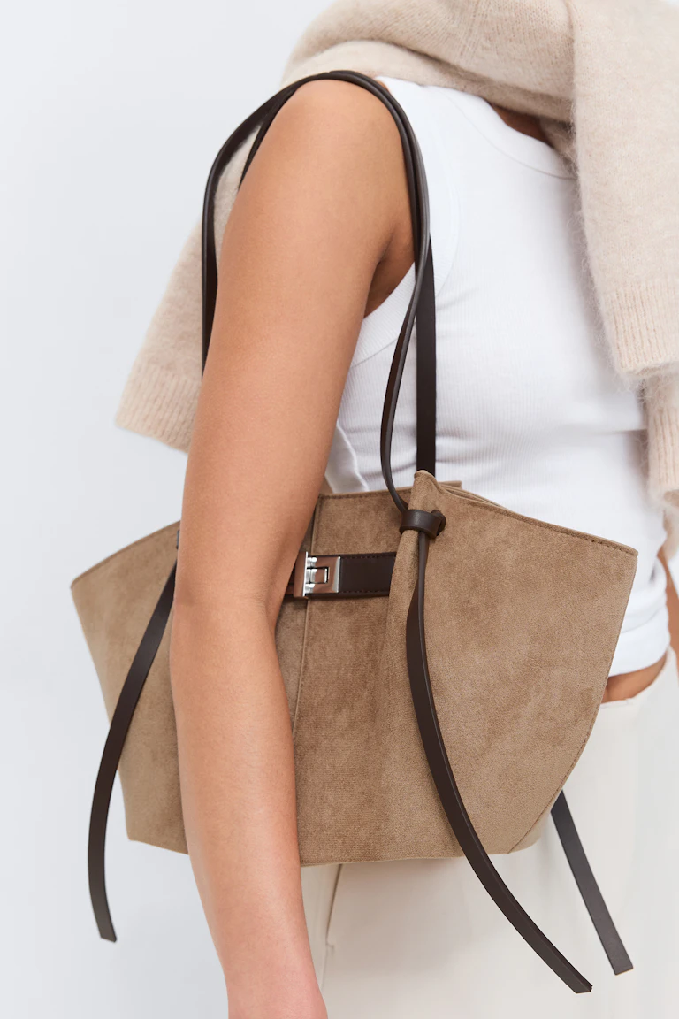 Witte geribde tanktop met een beige gebreide trui gedrapeerd over de schouders, crèmekleurige broek en een tan suède tote bag met donkerbruine leren schouderbanden en zilverkleurige gesp-hardware.