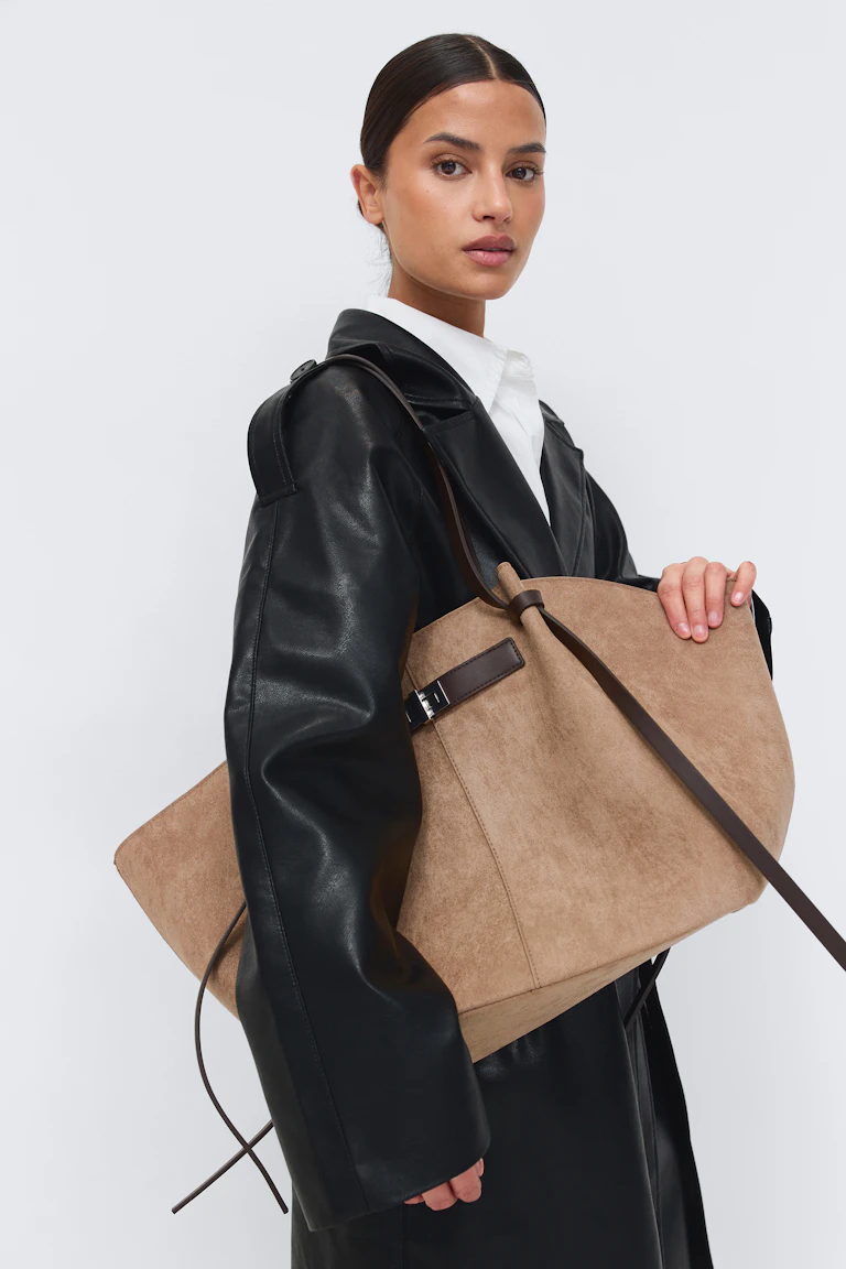 Trench-coat en cuir noir sur une chemise blanche à col, assorti d'un grand sac en daim fauve avec des lanières en cuir marron foncé et une boucle.