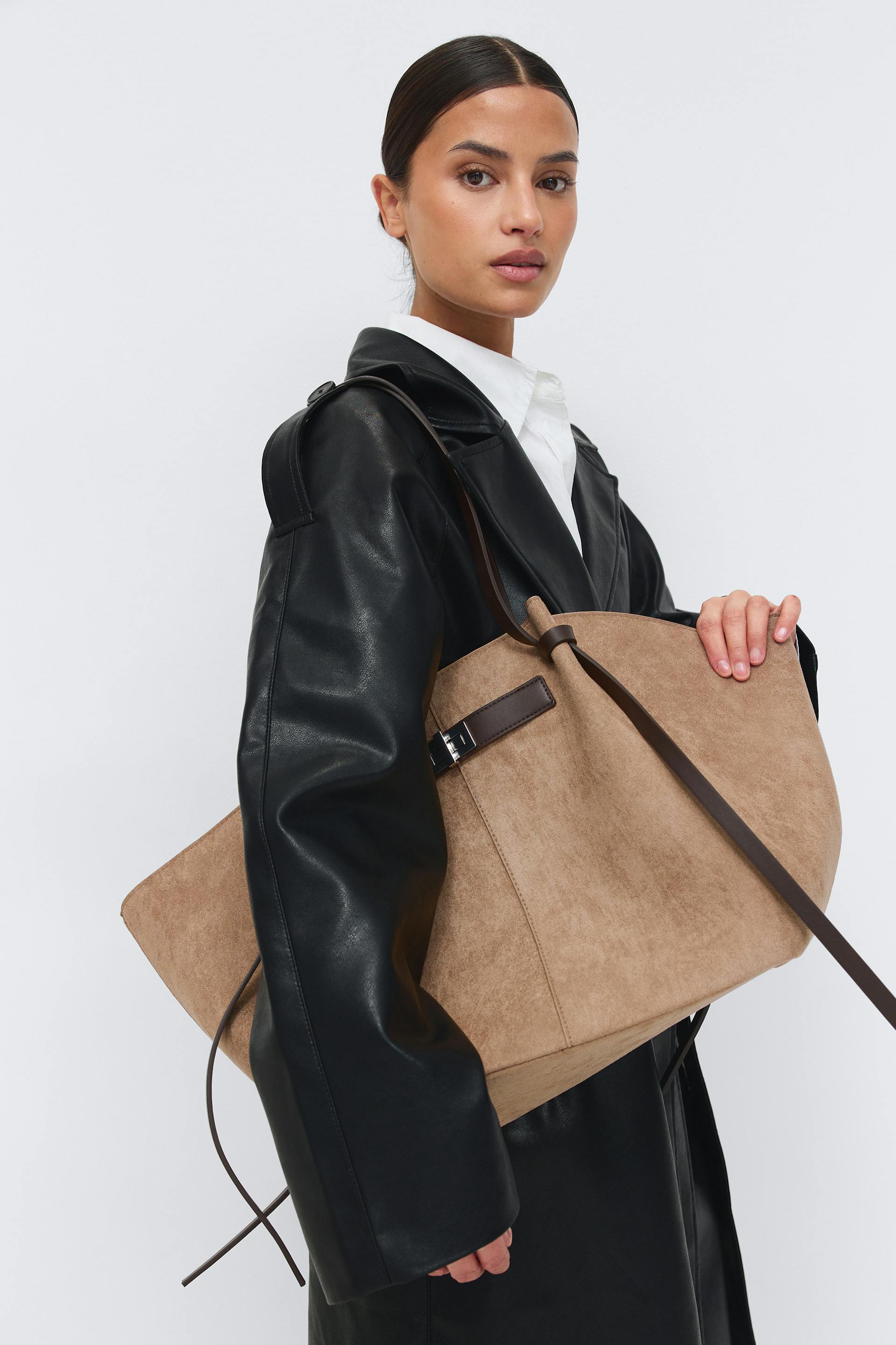 Black leather trench coat over a white collared shirt, paired with a large tan suede tote featuring dark brown leather straps and a buckle.