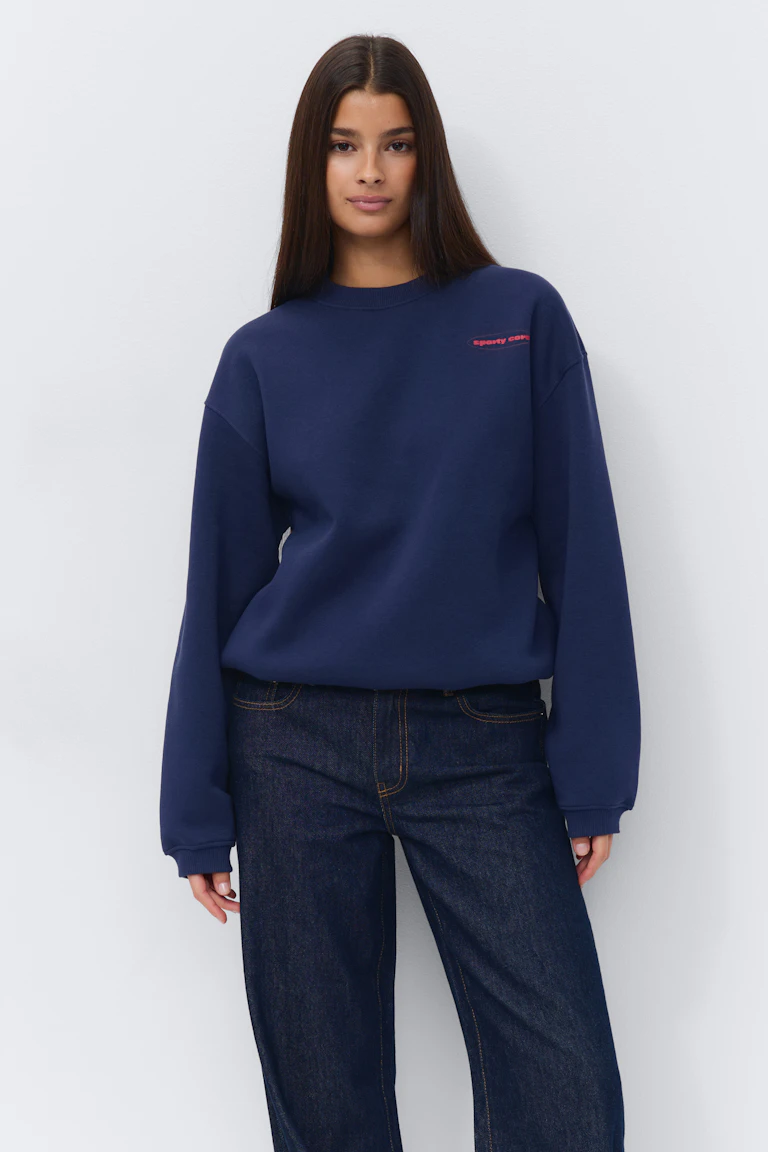 Navy crewneck sweater met een klein rood logo op de borst, gecombineerd met een donker indigo straight-leg jeans met contrasterende stiksels.