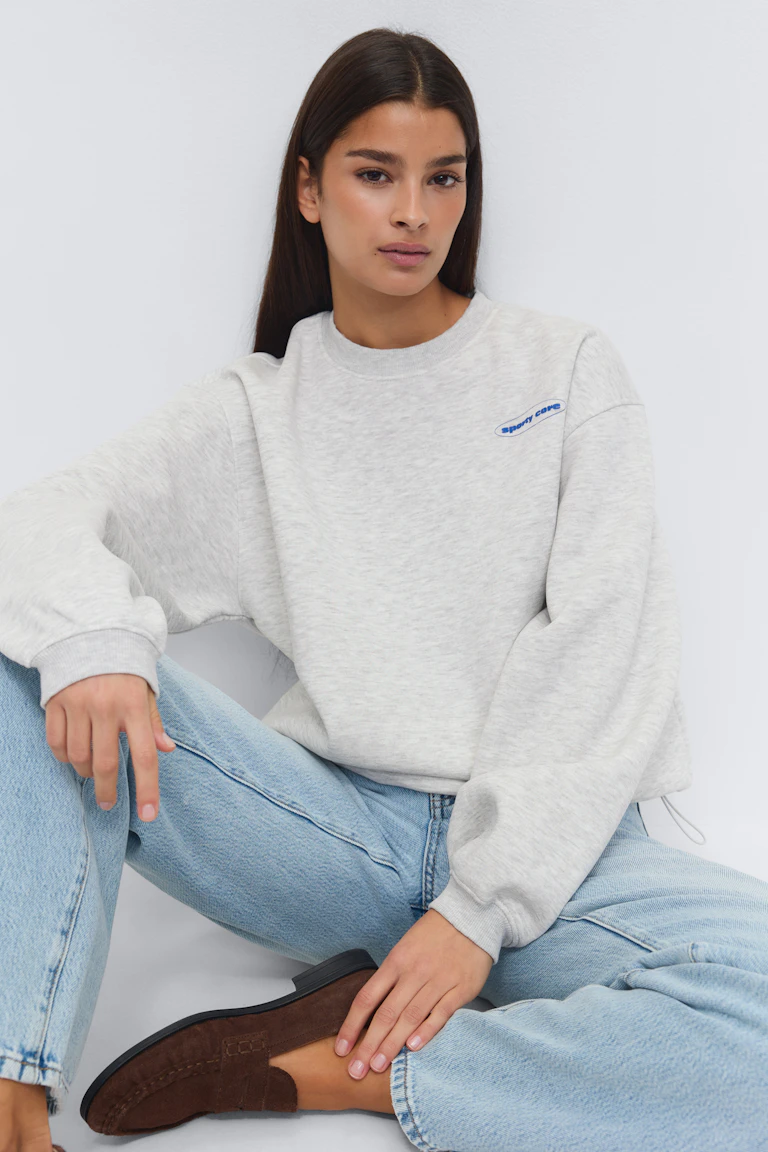 Helles, heidegraues Sweatshirt mit Rundhalsausschnitt, gerippten Bündchen und kleinem blauen, ovalen Logo auf der linken Brust; helle Jeans mit geradem Bein; dunkelbraune Wildleder-Slipper.