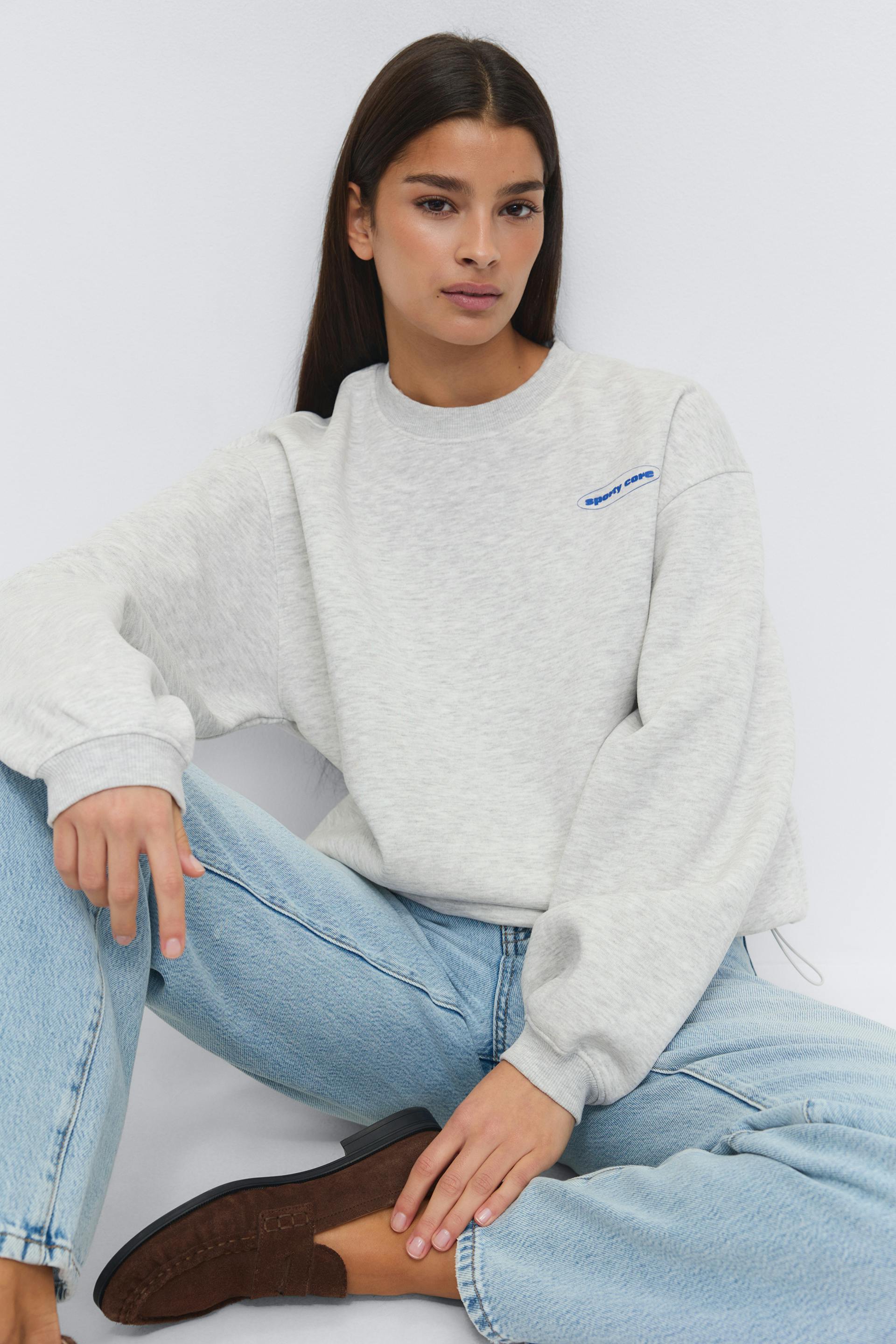 Light heather-gray crewneck sweatshirt with ribbed cuffs and small blue oval logo at left chest; light-wash straight-leg jeans; dark brown suede loafers.