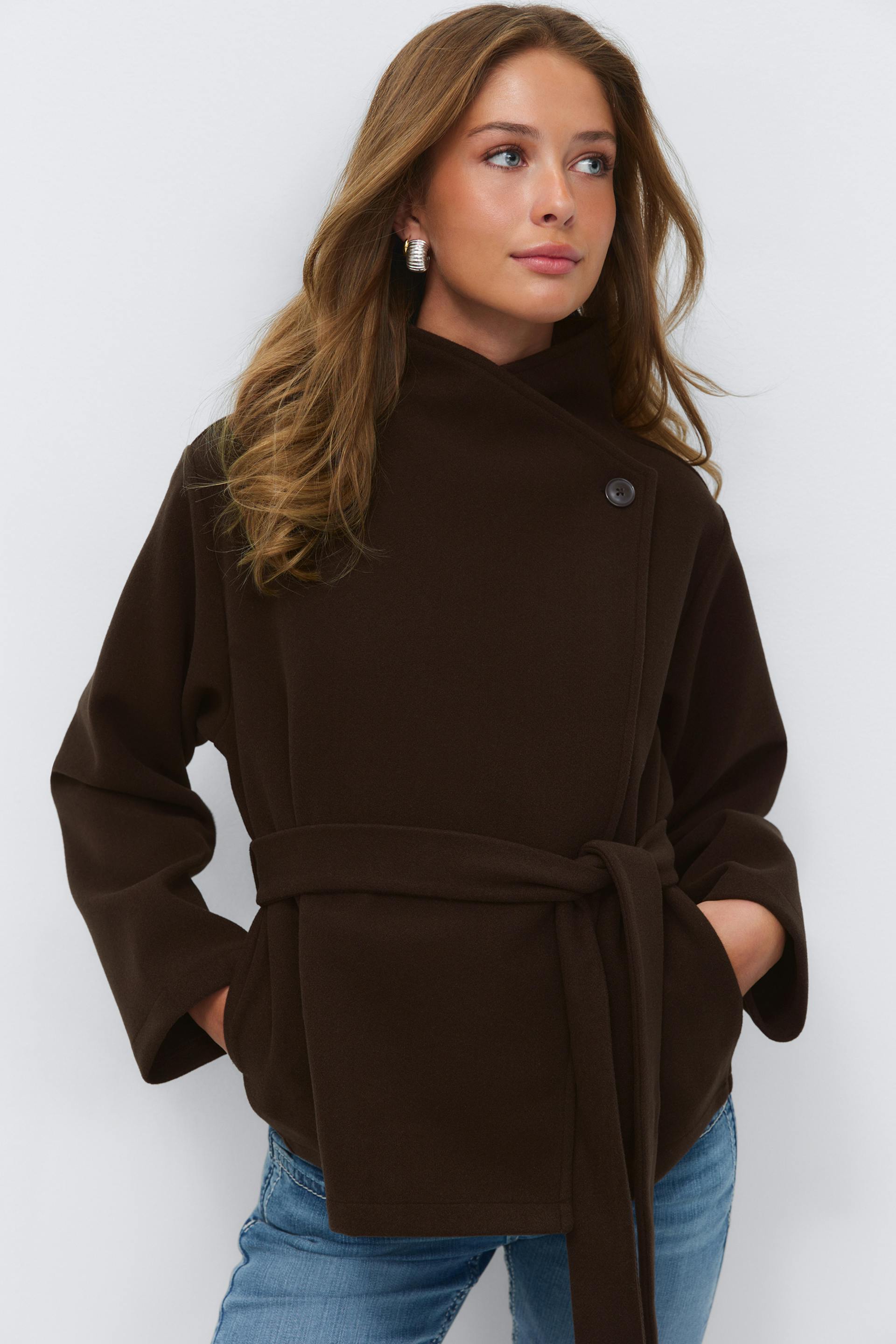 Dark brown belted wrap coat with a high asymmetrical collar and single-button detail, paired with light-blue denim jeans.