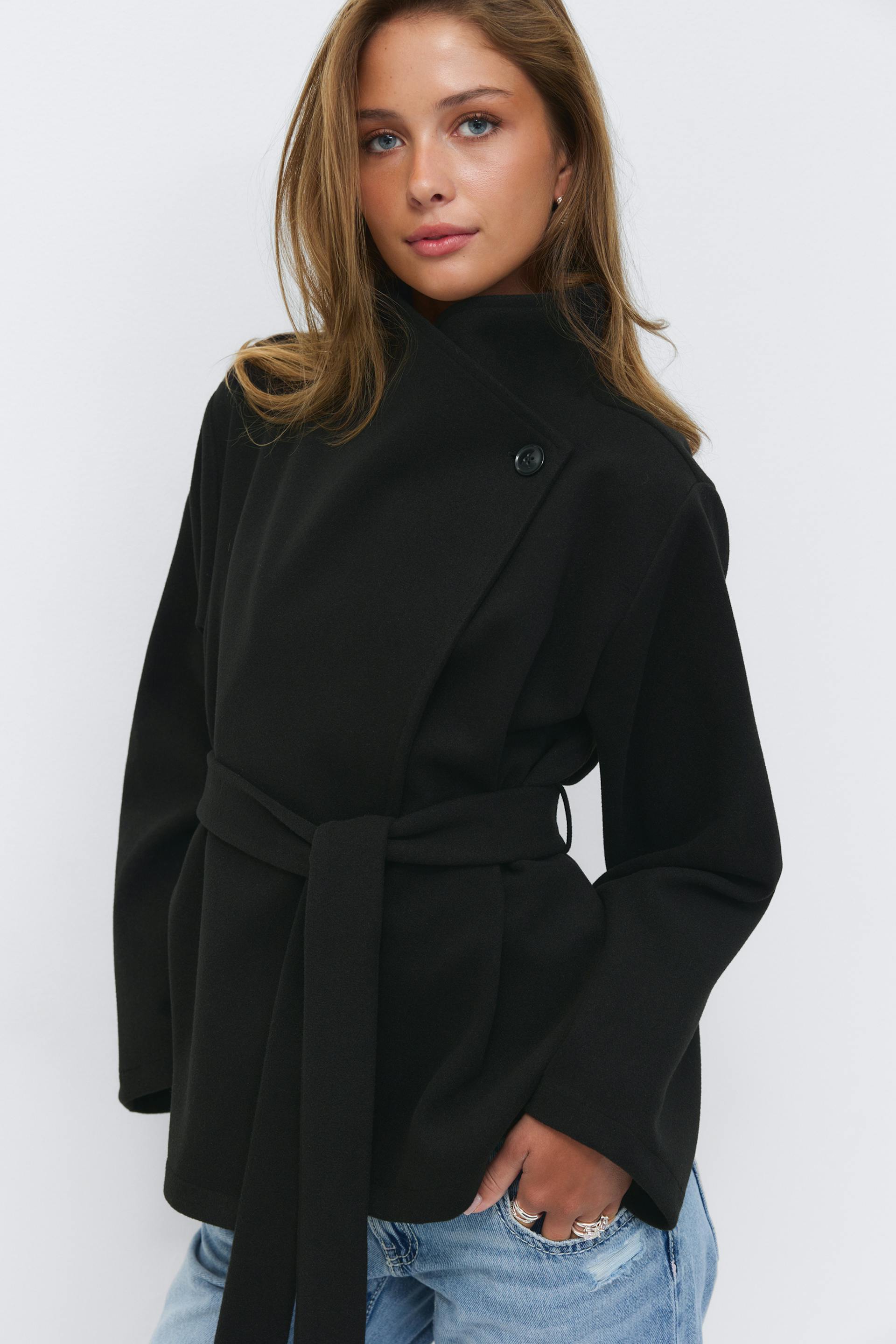 Black belted wrap coat with an asymmetric high collar and single-button detail, worn with light-wash distressed jeans.