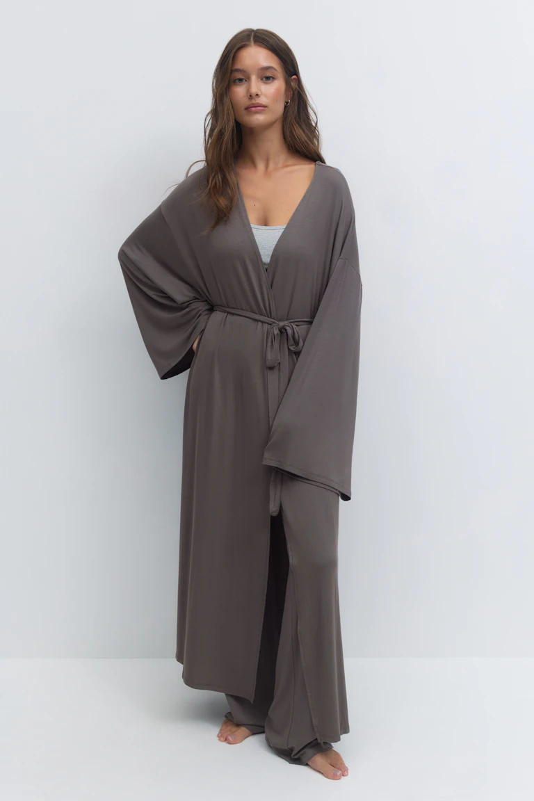 Frau trägt ein langes taupe-graues Gewand im Kimono-Stil mit weiten Ärmeln und einem Gürtel zum Binden in der Taille, darüber ein hellgraues Unterhemd und eine passende Lounge-Hose mit weitem Bein, barfuß.