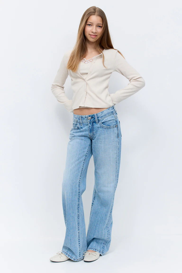 Beige langærmet cropped cardigan med knapper over en blondekantet camisole, lysvaskede lavtaljede jeans med vide ben og cremefarvede ballerinaer med sløjfer.