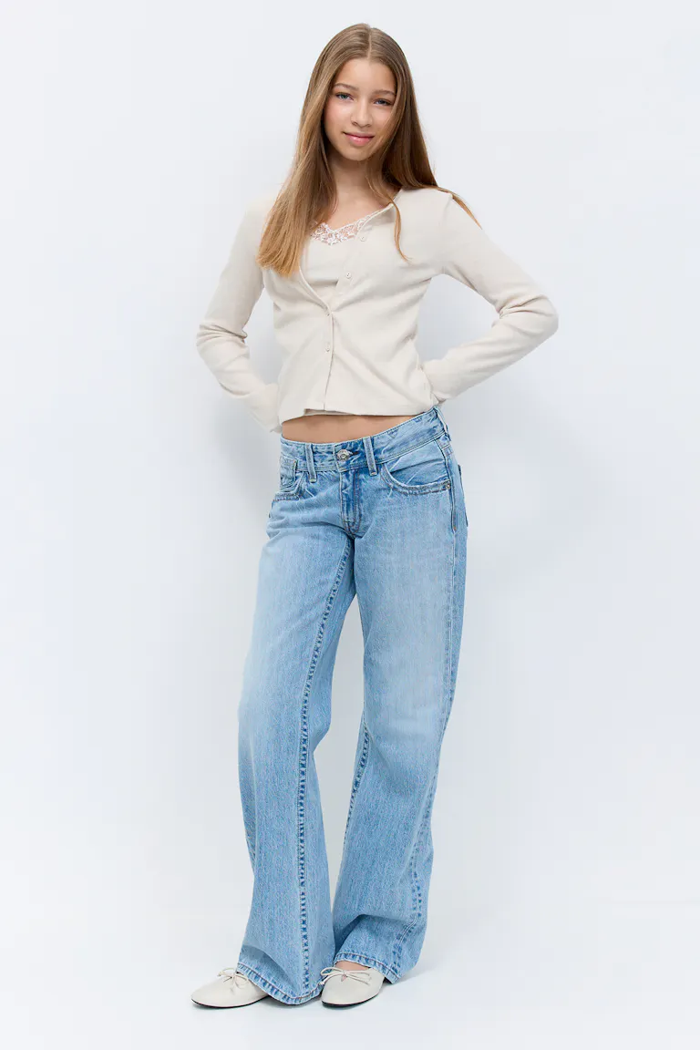 Beige langærmet cropped cardigan med knapper over en blondekantet camisole, lysvaskede lavtaljede jeans med vide ben og cremefarvede ballerinaer med sløjfer.