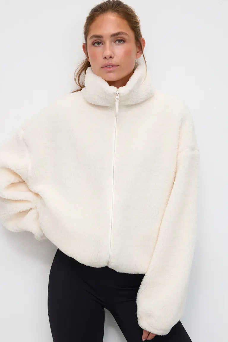Ivory sherpa-fleece zip-up jacket with a high funnel collar and voluminous long sleeves, styled with black fitted leggings.