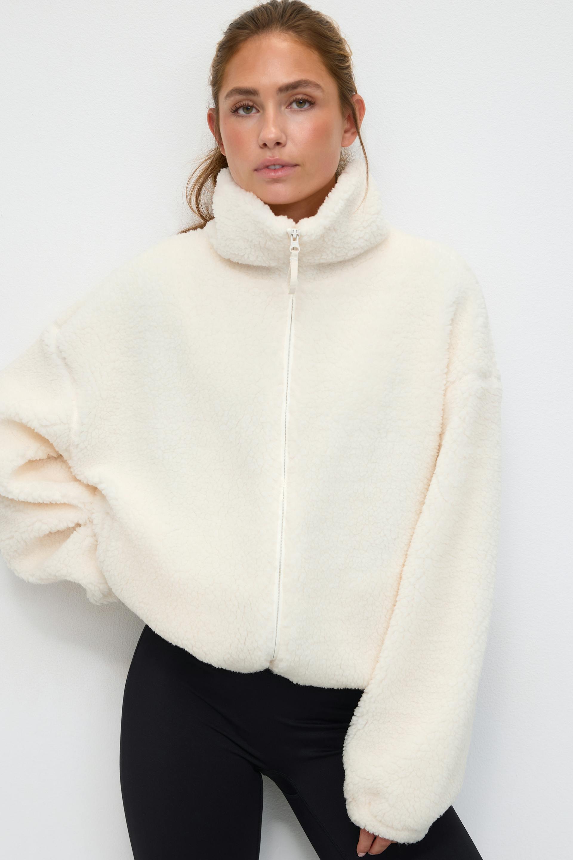 Elfenbeinfarbene Sherpa-Fleece-Jacke mit Reißverschluss, hohem Trichterkragen und voluminösen, langen Ärmeln, kombiniert mit schwarzen, taillierten Leggings.