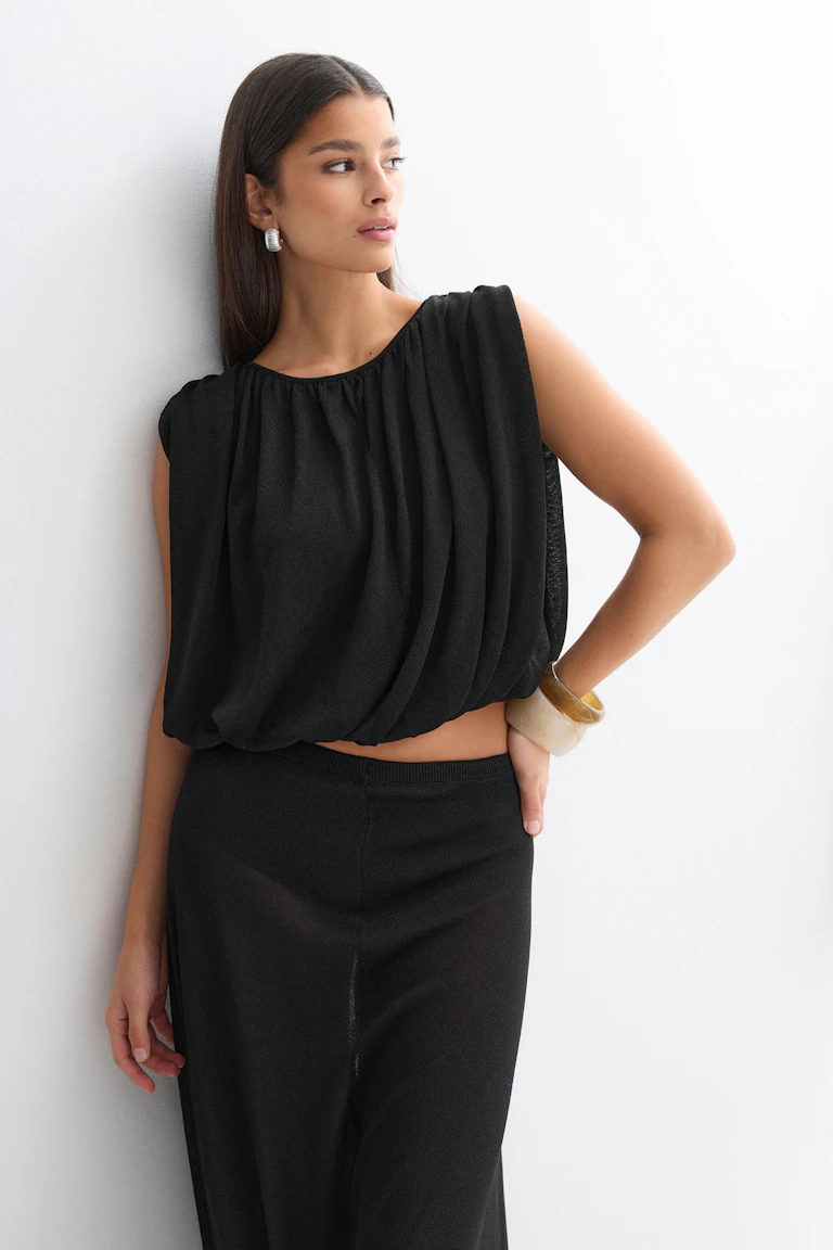 Svart ärmlös draperad crop top med rund halsringning kombinerad med en svart kjol med hög midja, stylad med silverfärgade örhängen och staplade armband i harts.