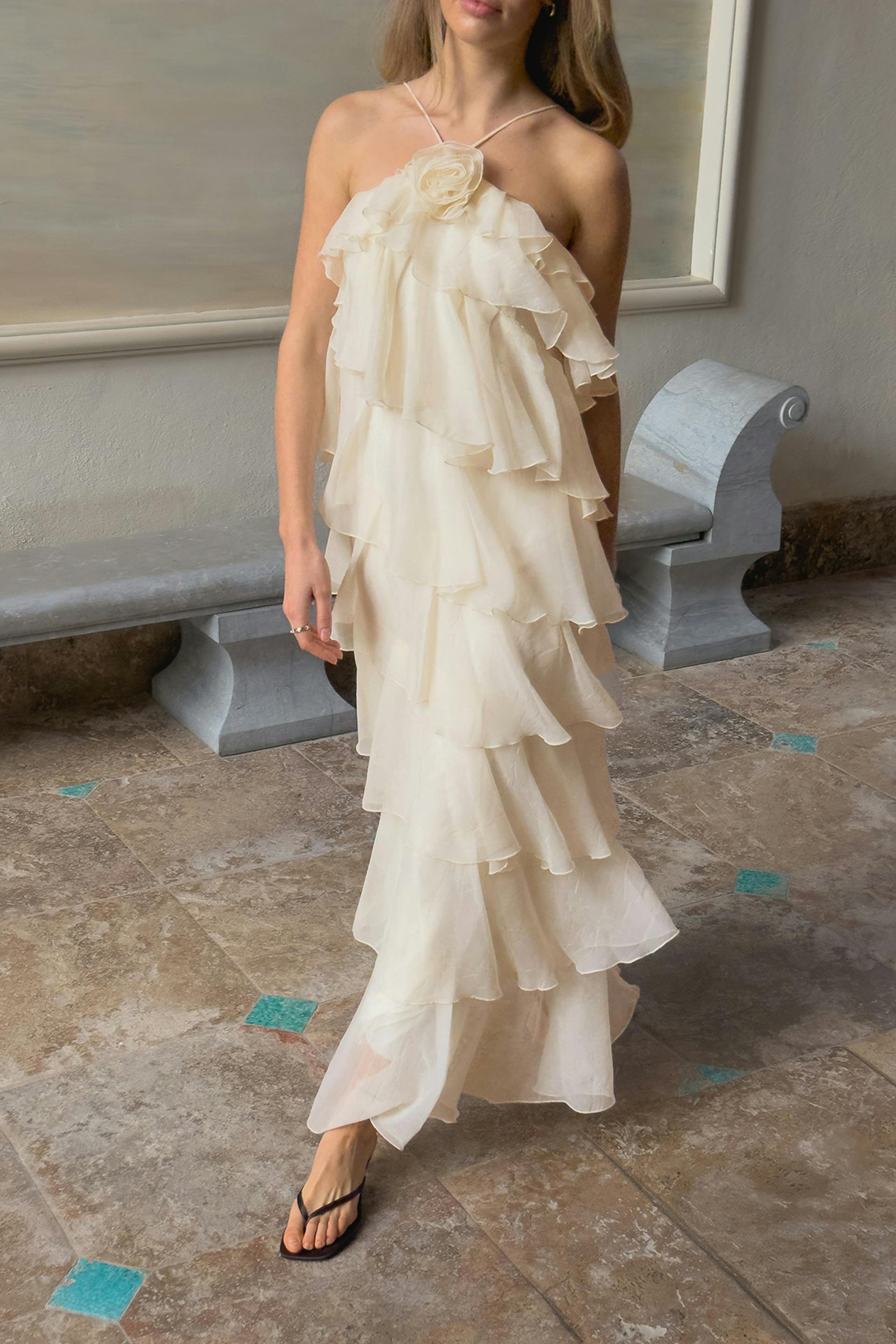 Norsunluunvärinen porrastettu sifonkinen halter-mekko, jossa on ryöppyäviä röyhelöitä ja kukka-applikaatio kaula-aukolla, yhdistettynä mustiin tangasandaaleihin.