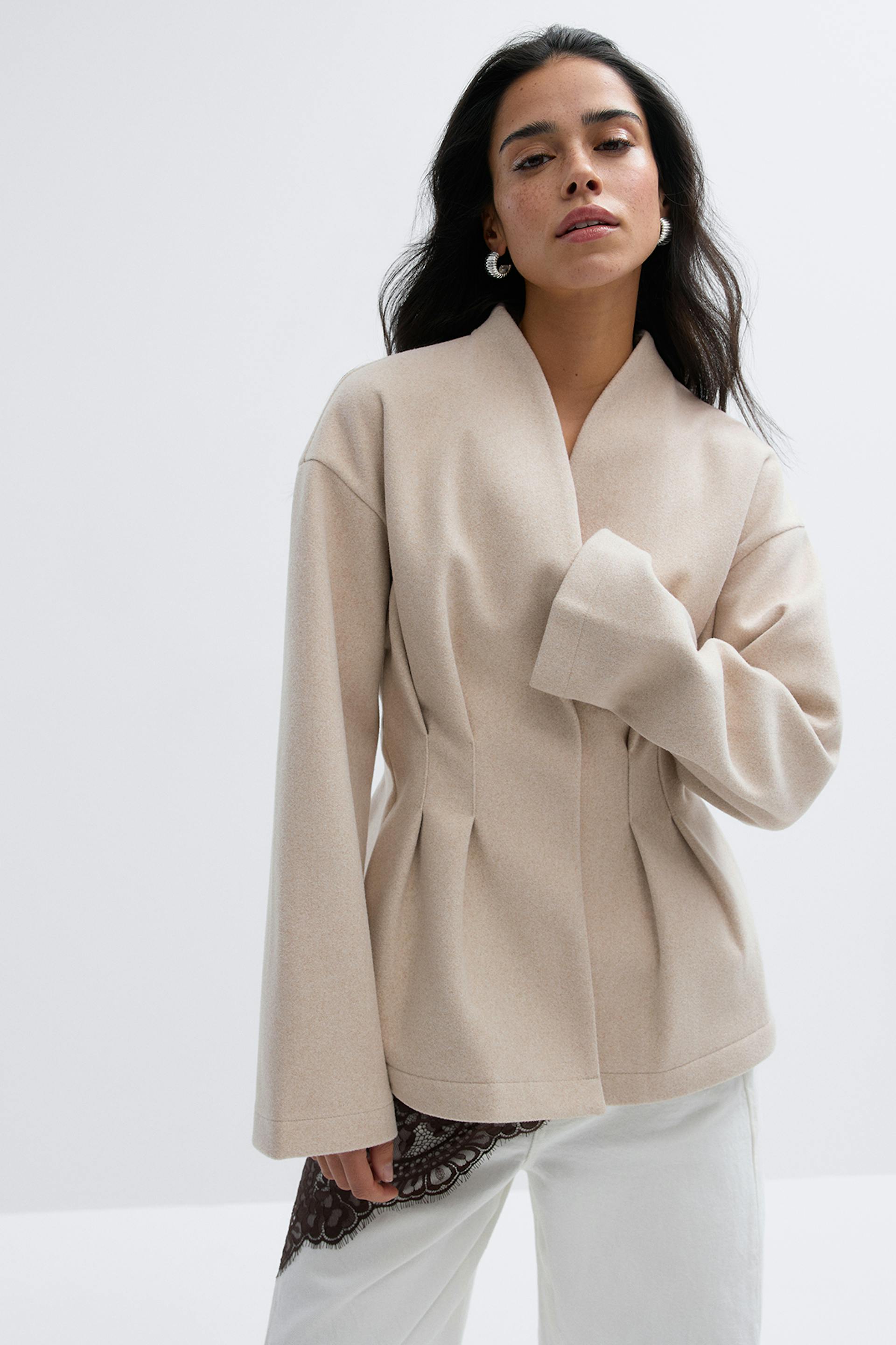 Beige räätälöity villatakki, jossa on huivikaulus, pitkät levenevät hihat ja vyötärön laskokset, yhdistettynä valkoisiin housuihin, joissa on musta pitsikoriste.