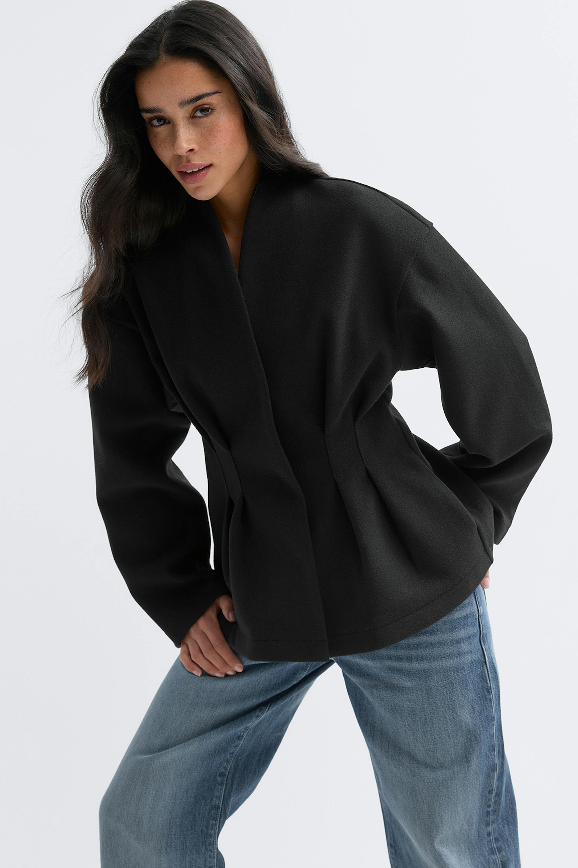 Musta strukturoitu takki, jossa on hienovarainen huivikaulus, pitkät hihat ja räätälöidyt vyötärön laskokset, yhdistettynä vaaleaksi pestyihin suoralahkeisiin farkkuihin.