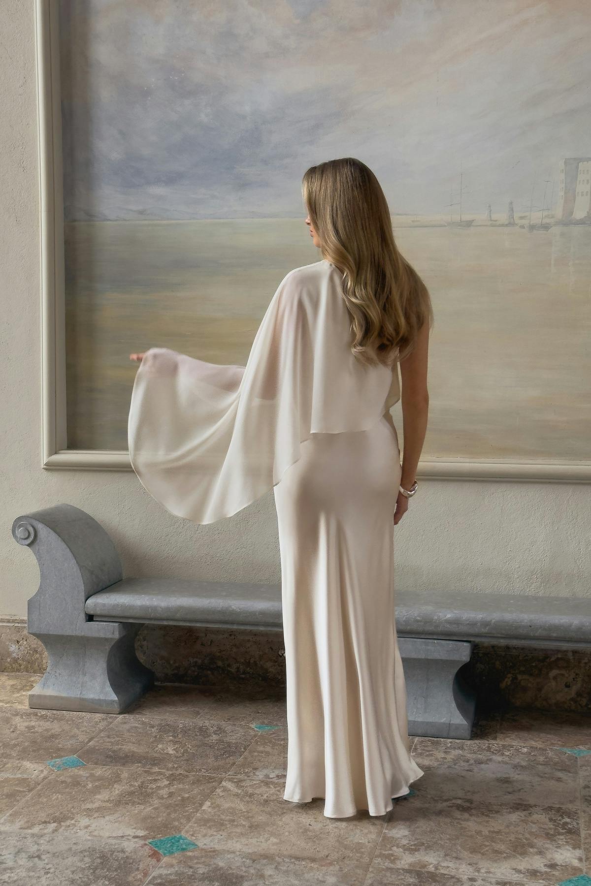 Lattianmyötäinen norsunluunvärinen satiinipuku, jossa on istuva siluetti ja epäsymmetrinen silkkinen rapattu viitta toisella olkapäällä.