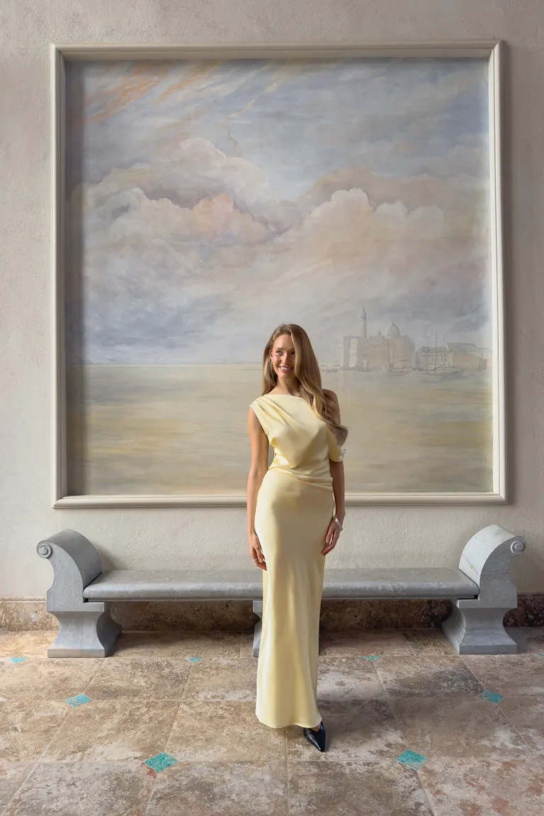 Pale yellow satin asymmetrical draped floor-length gown with a fitted waist, paired with black pointed-toe heels and a wristwatch.
