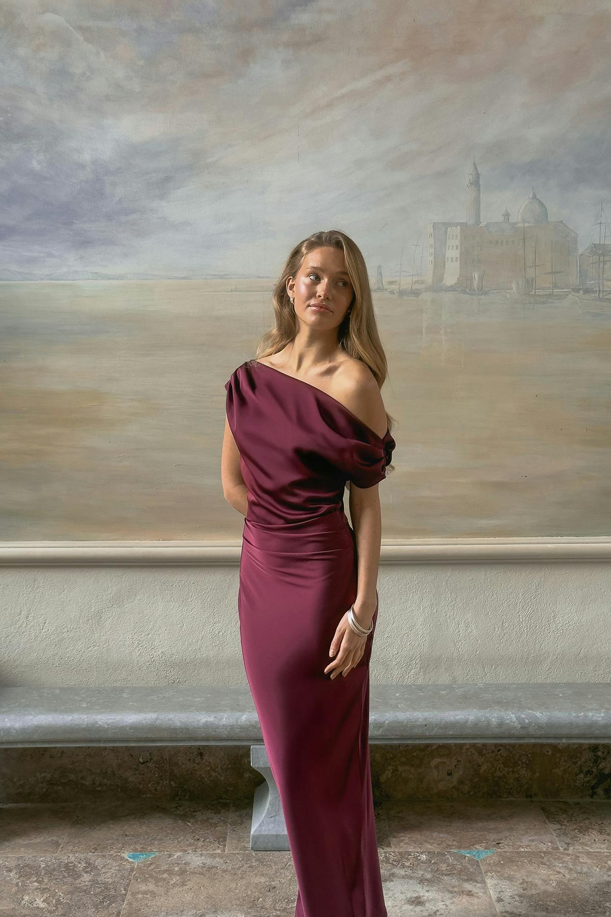 Lattianmyötäinen syvän viininpunainen yhden olkapään satiini iltapuku, jossa on rapattu off-shoulder-hihansuojus ja istuva vyötärö; koristeltu ohuilla metallisilla rannerenkailla ja pienillä vanne-korvakoruilla.