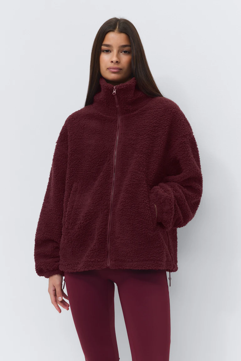 Burgunderfarget sherpajakke med glidelås, høy traktkrage og sidelommer, kombinert med figursydde burgunderfargede leggings.