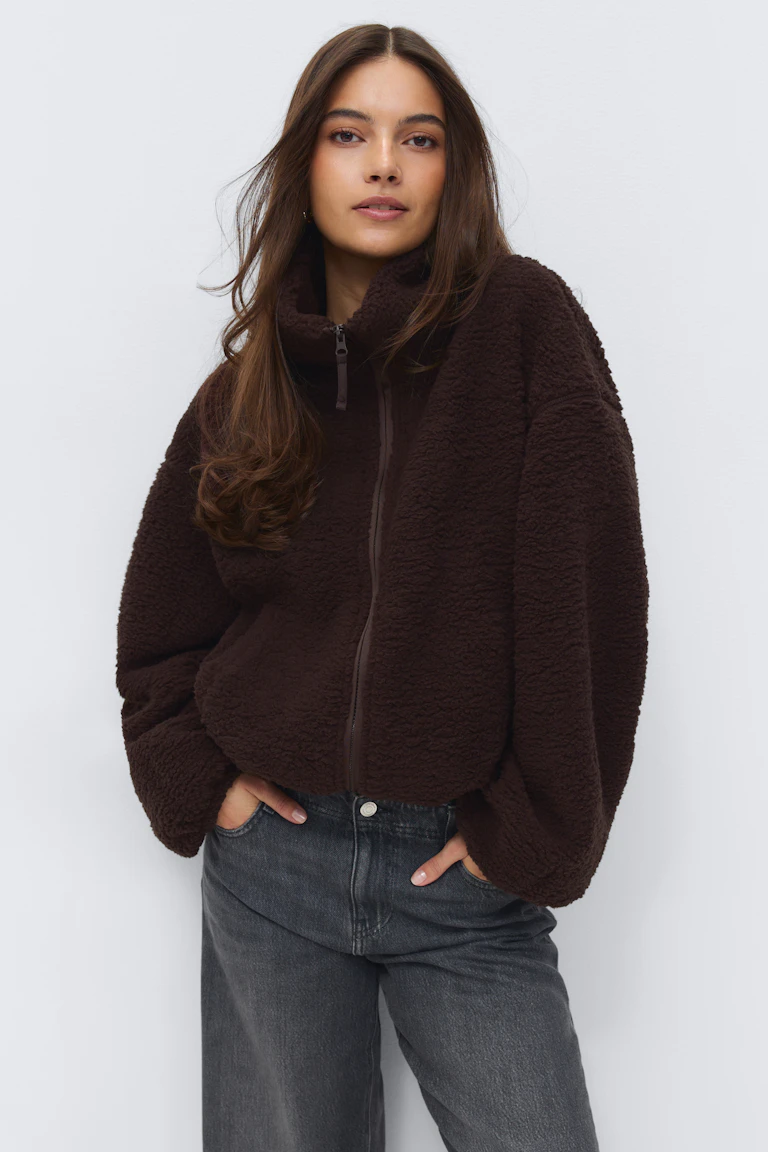 Veste zippée en sherpa marron foncé avec col cheminée et manches volumineuses, associée à un jean taille haute délavé gris noir.