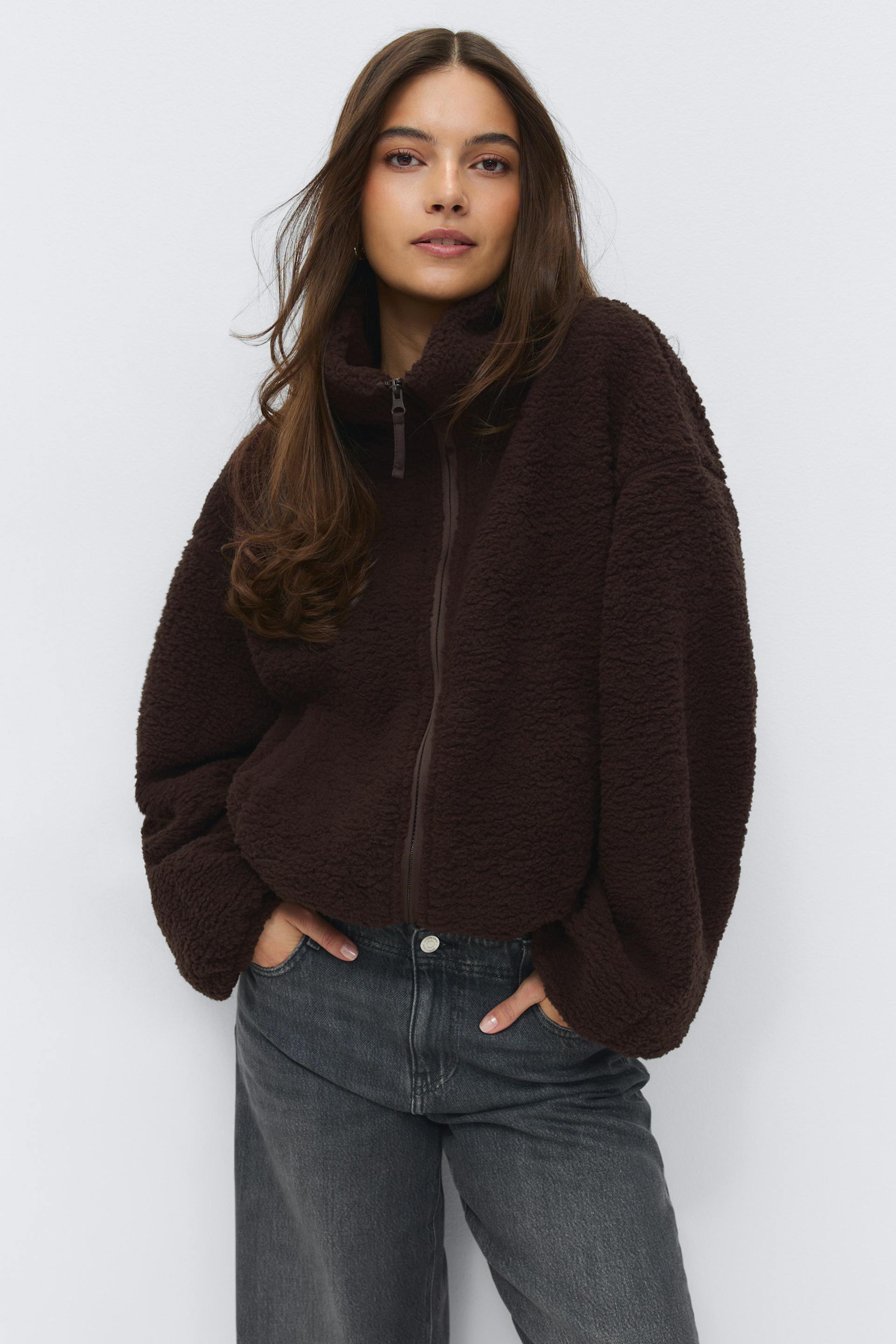 Dark brown sherpa zip-up jacket with high funnel collar and voluminous sleeves, paired with high‑waisted faded black‑grey jeans.