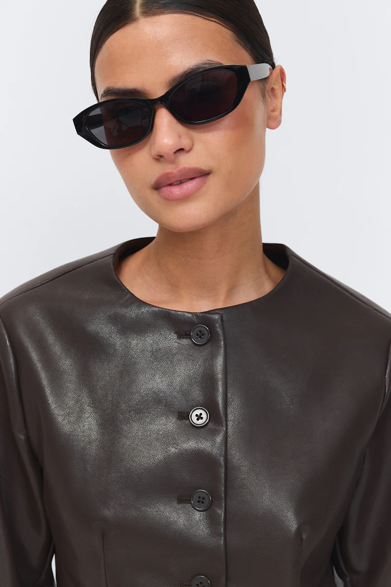 Black cat‑eye sunglasses and a dark brown leather button‑front jacket with a rounded neckline.
