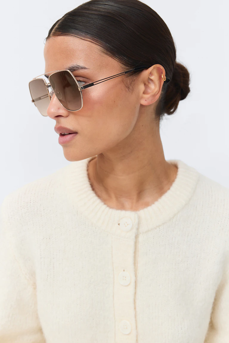 Elfenbensfarget strikket cardigan med ribbestrikket rund hals og tonede knapper, kombinert med overdimensjonerte, firkantede solbriller med gradientglass, tynn gullramme og svarte brillestenger.