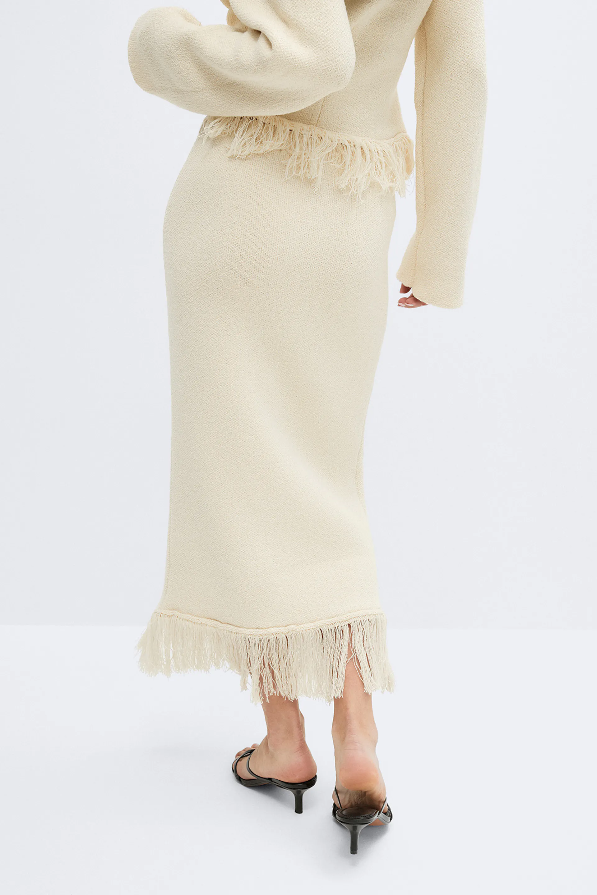 Cremefarbener, strukturierter Strick-Midirock mit Fransensaum, kombiniert mit einer passenden, kurzen Jacke mit Fransenbesatz und schwarzen Pantoletten mit niedrigem Absatz.