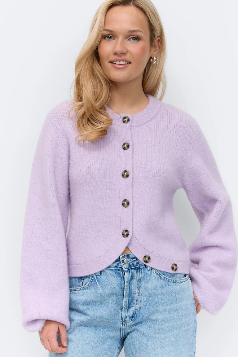 Lilla cardigan med rund hals og knapper i skilpaddeskinn, buet kant i nederkant og lange ermer med litt ballongformede former, kombinert med lyse, høytaljede jeans.