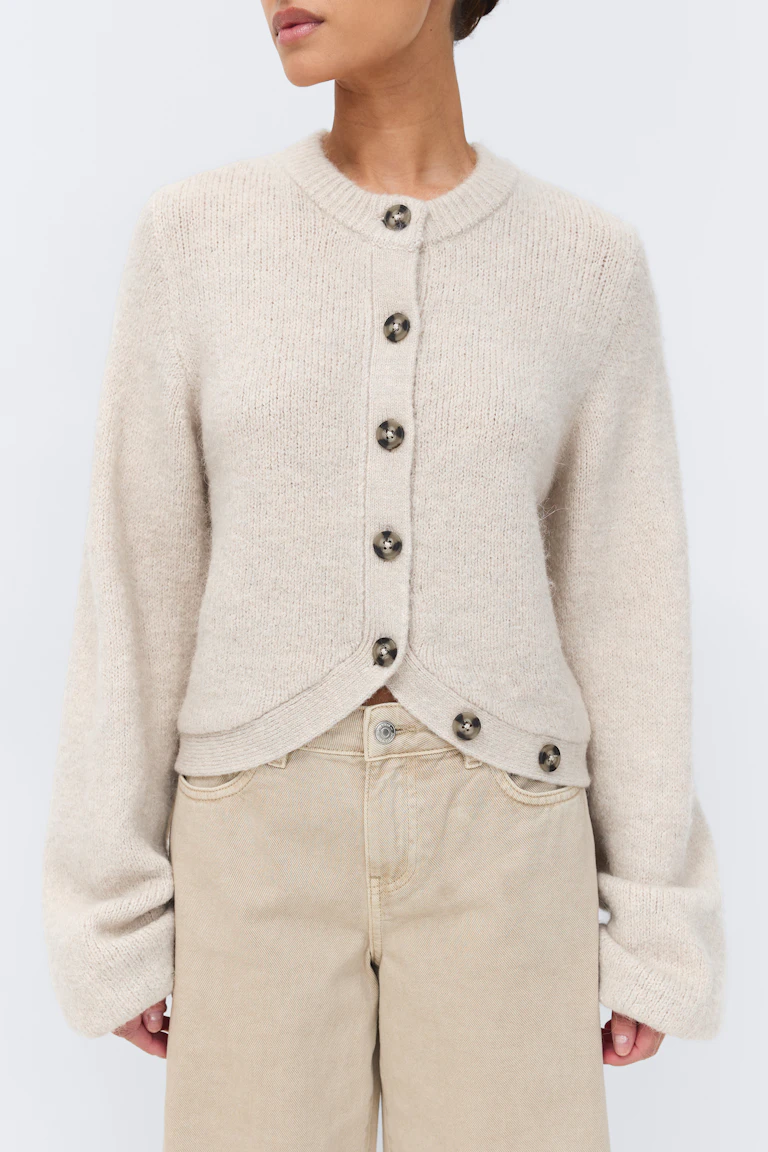 Lichtbeige cardigan met ronde hals en knoopsluiting, grote schildpadknopen, afgeronde zoom en lange ontspannen mouwen, gecombineerd met een beige broek met hoge taille.