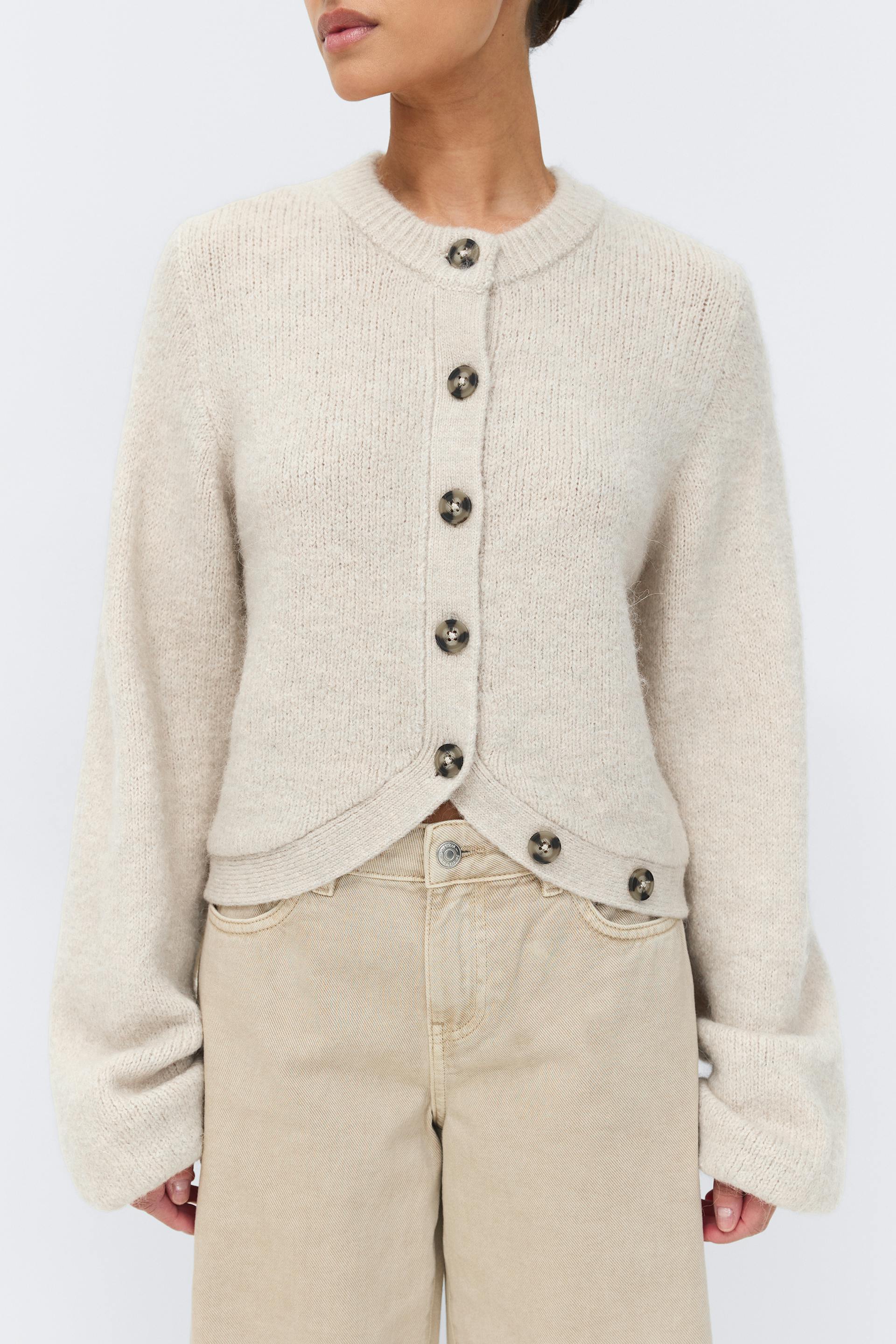 Light beige crew‑neck button‑front cardigan with large tortoiseshell buttons, curved cropped hem and long relaxed sleeves, paired with high‑waisted beige trousers.