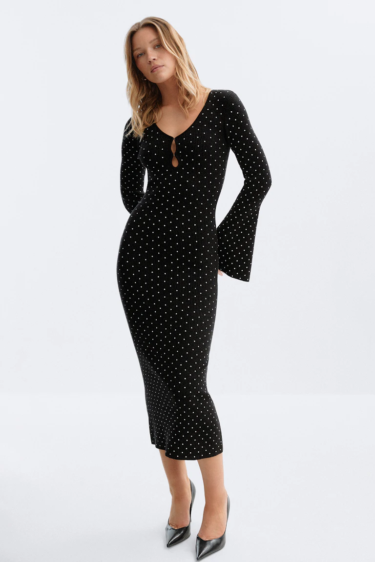 Robe midi noire en maille côtelée à pois blancs, col en V avec deux découpes en trou de serrure et manches longues évasées ; associée à des escarpins noirs à bouts pointus.