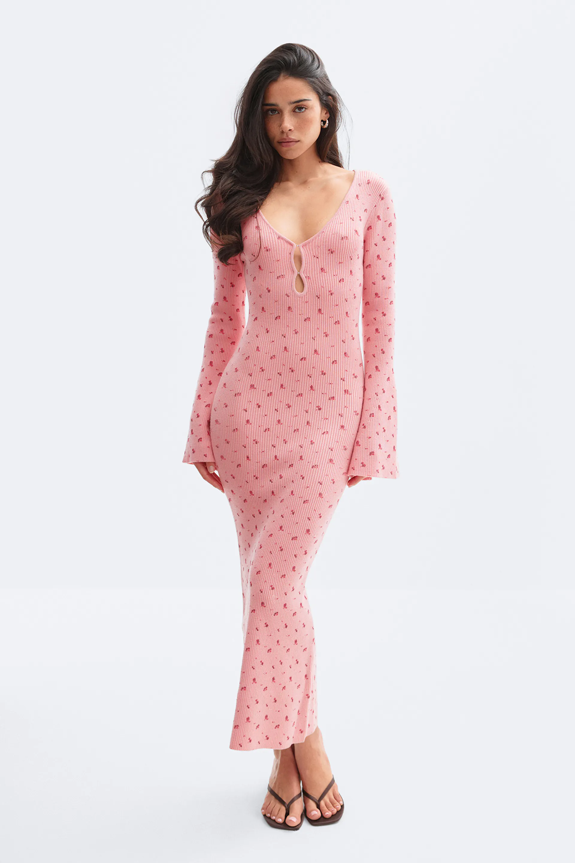 Rosa, figurbetontes Maxikleid aus Rippstrick mit langen, ausgestellten Ärmeln, tiefem V-Ausschnitt mit zwei Schlüssellochausschnitten und kleinem Blumenmuster, kombiniert mit schwarzen Zehensandalen.