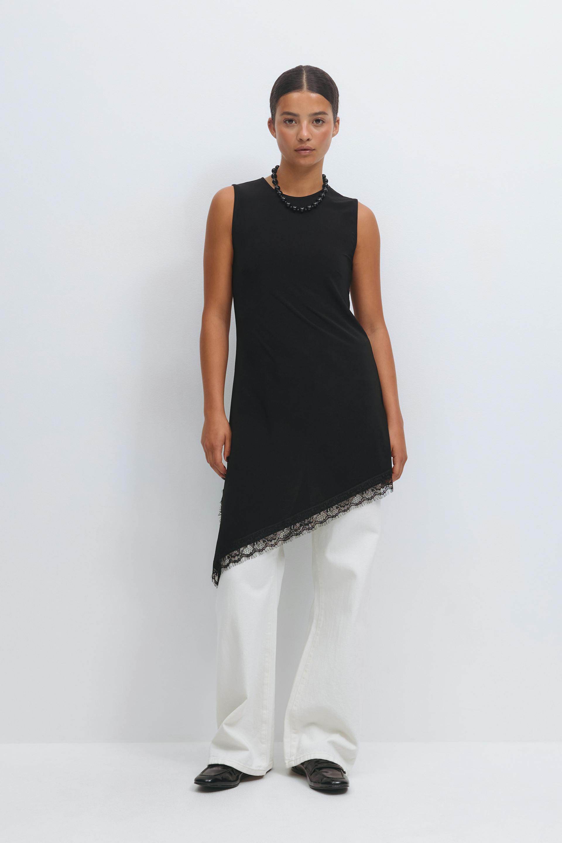 Sleeveless black asymmetrical tunic with lace-trimmed diagonal hem, paired with white wide-leg trousers, black loafers and a short black beaded necklace.