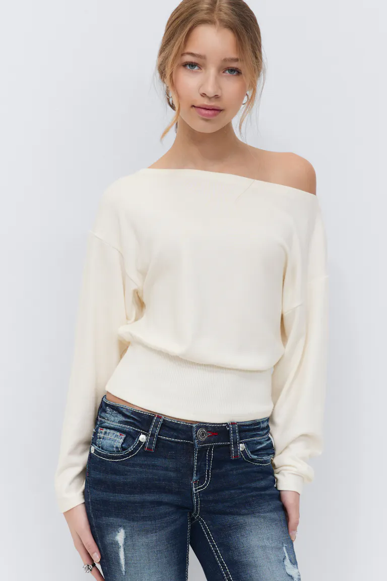 Kermanvärinen pitkähihainen off-the-shoulder-pusero, jossa on uurrettu vyötärönauha, yhdistettynä tummaksi pestyihin distressed-farkkuihin, joissa on valkoiset kontrastiompeleet.