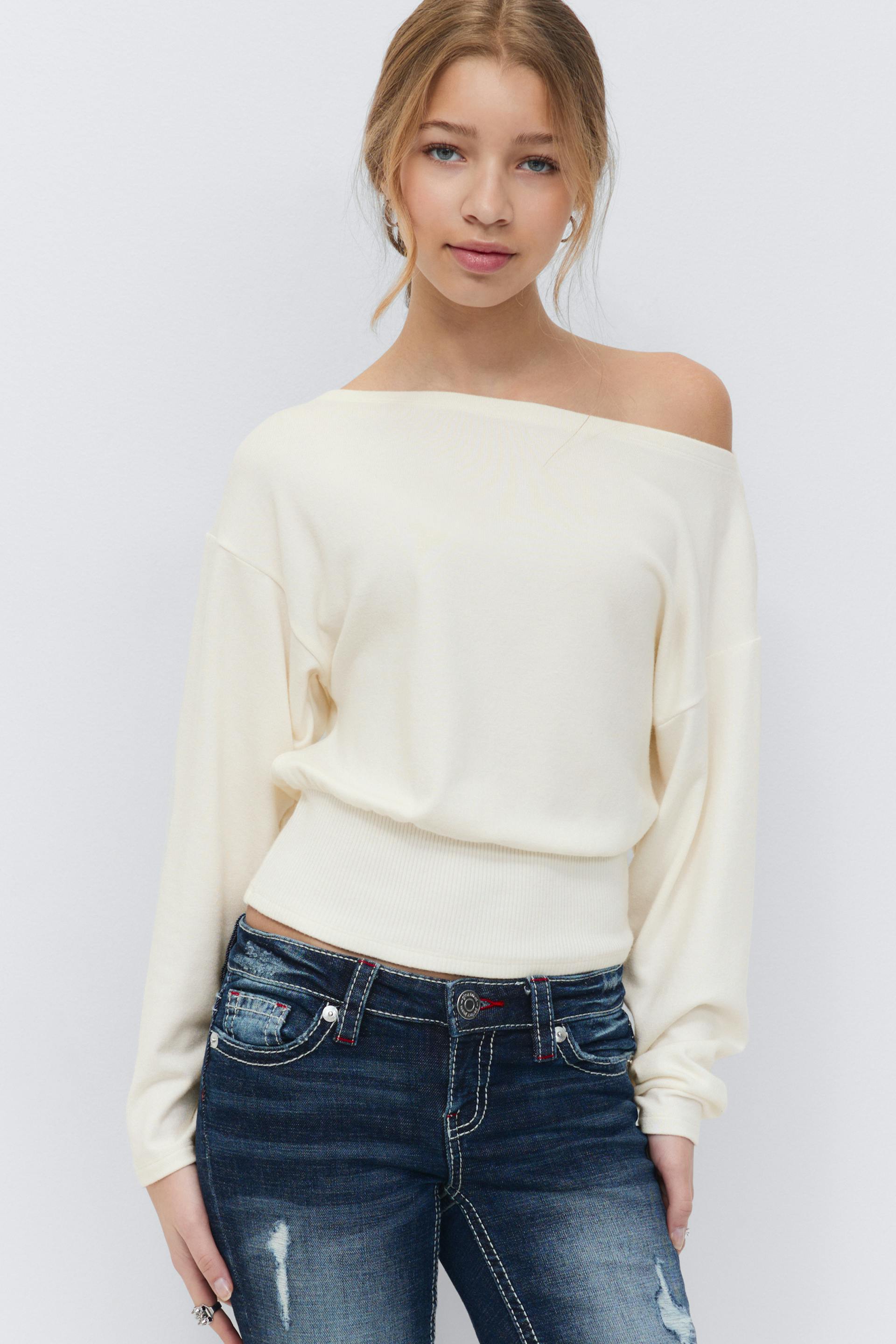 Kermanvärinen pitkähihainen off-the-shoulder-pusero, jossa on uurrettu vyötärönauha, yhdistettynä tummaksi pestyihin distressed-farkkuihin, joissa on valkoiset kontrastiompeleet.