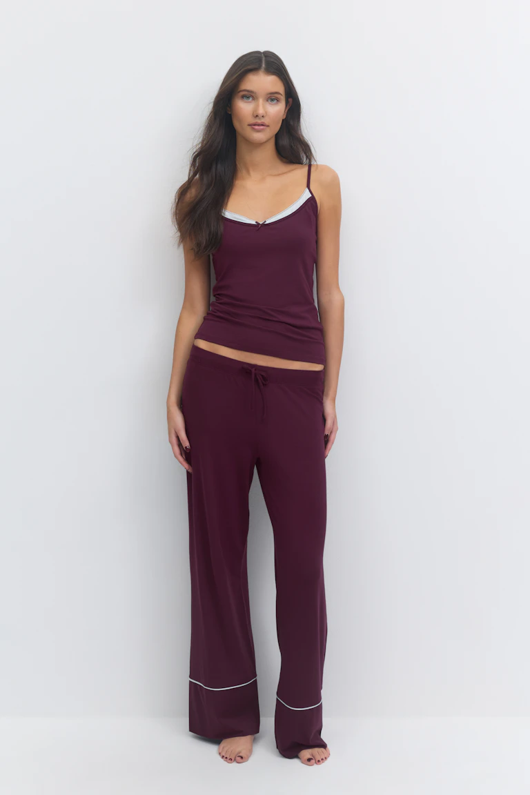 Syvä viininpunainen camisole, jossa on ohuet olkaimet, valkoisella reunustettu V-kaula-aukko ja pieni keskellä oleva rusetti, yhdistettynä yhteensopiviin leveälahkeisiin leveälahkeisiin oleskeluhousuihin, joissa on valkoinen putkitus reunojen yläpuolella.